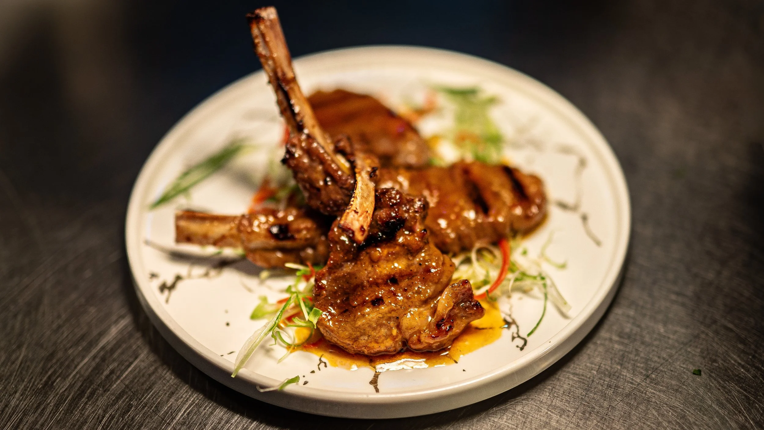 Lamb chops served on a white plate with greens and sauce, placed on a dark surface.
