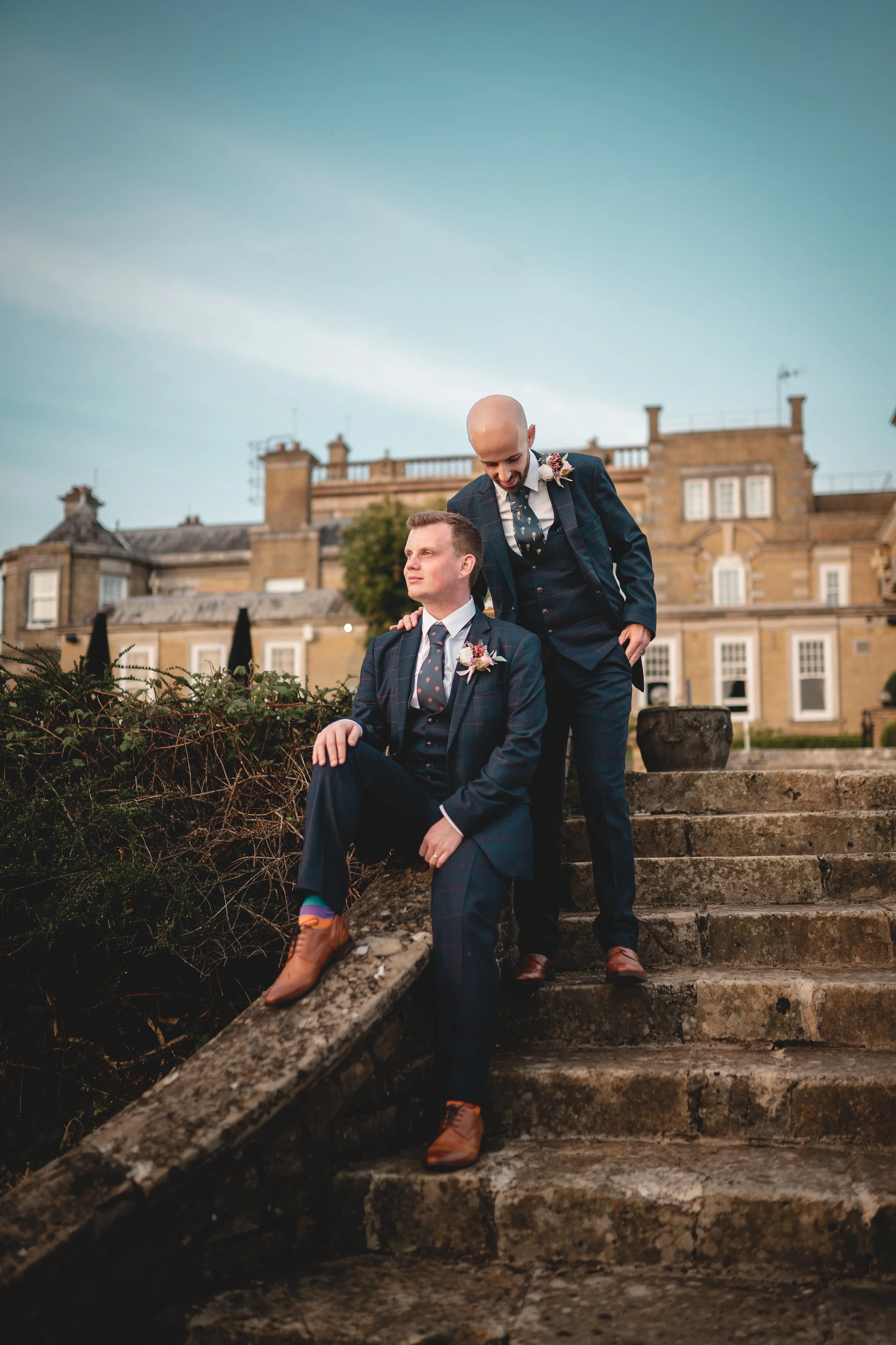 Two men dressed in suits, one seated and one with his hand on the other's shoulder, on outdoor stone steps with an old building in the background at sunset.