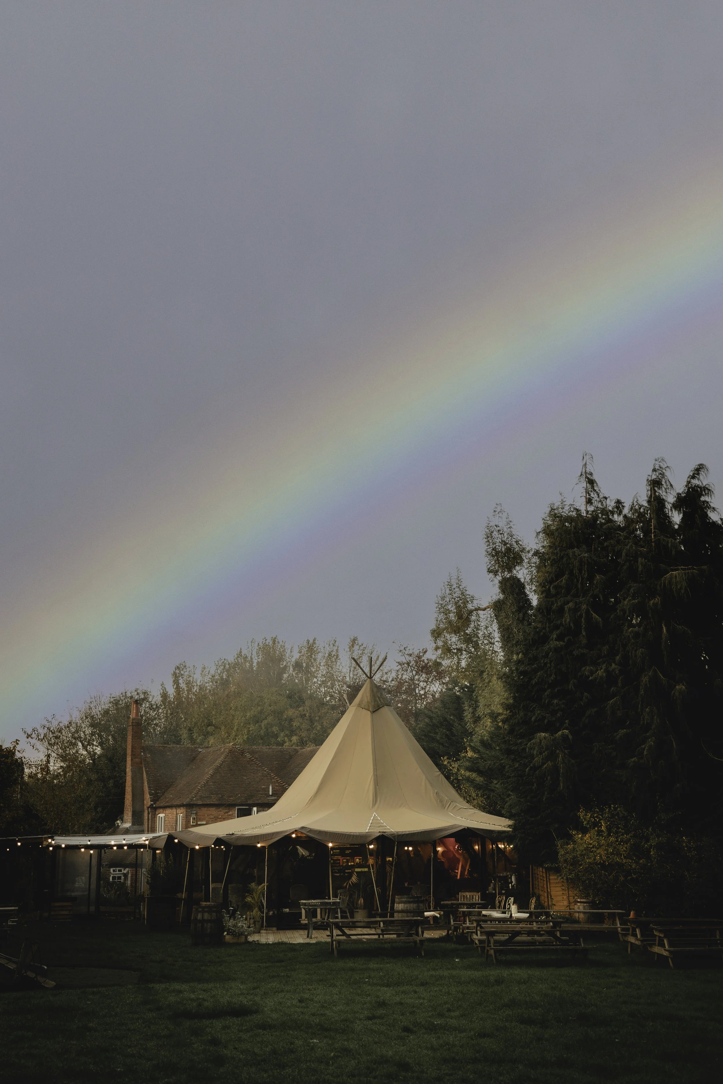 A rainbow in the sky over a backyard with a large beige tent, string lights, and outdoor furniture, with trees and houses in the background.