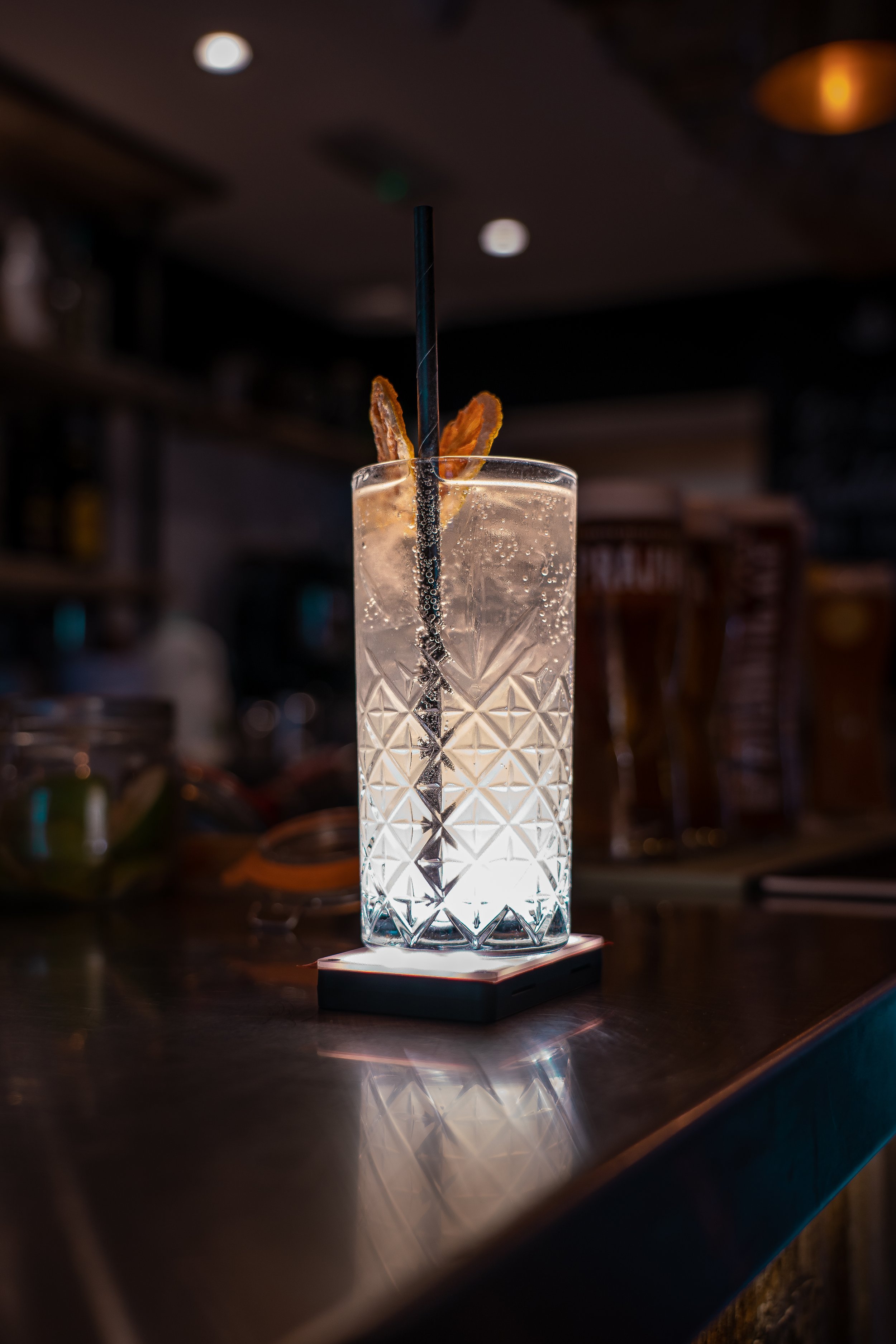 A tall glass of clear cocktail with bubbles, garnished with two slices of orange peel and a black straw, placed on a coaster on a bar counter.