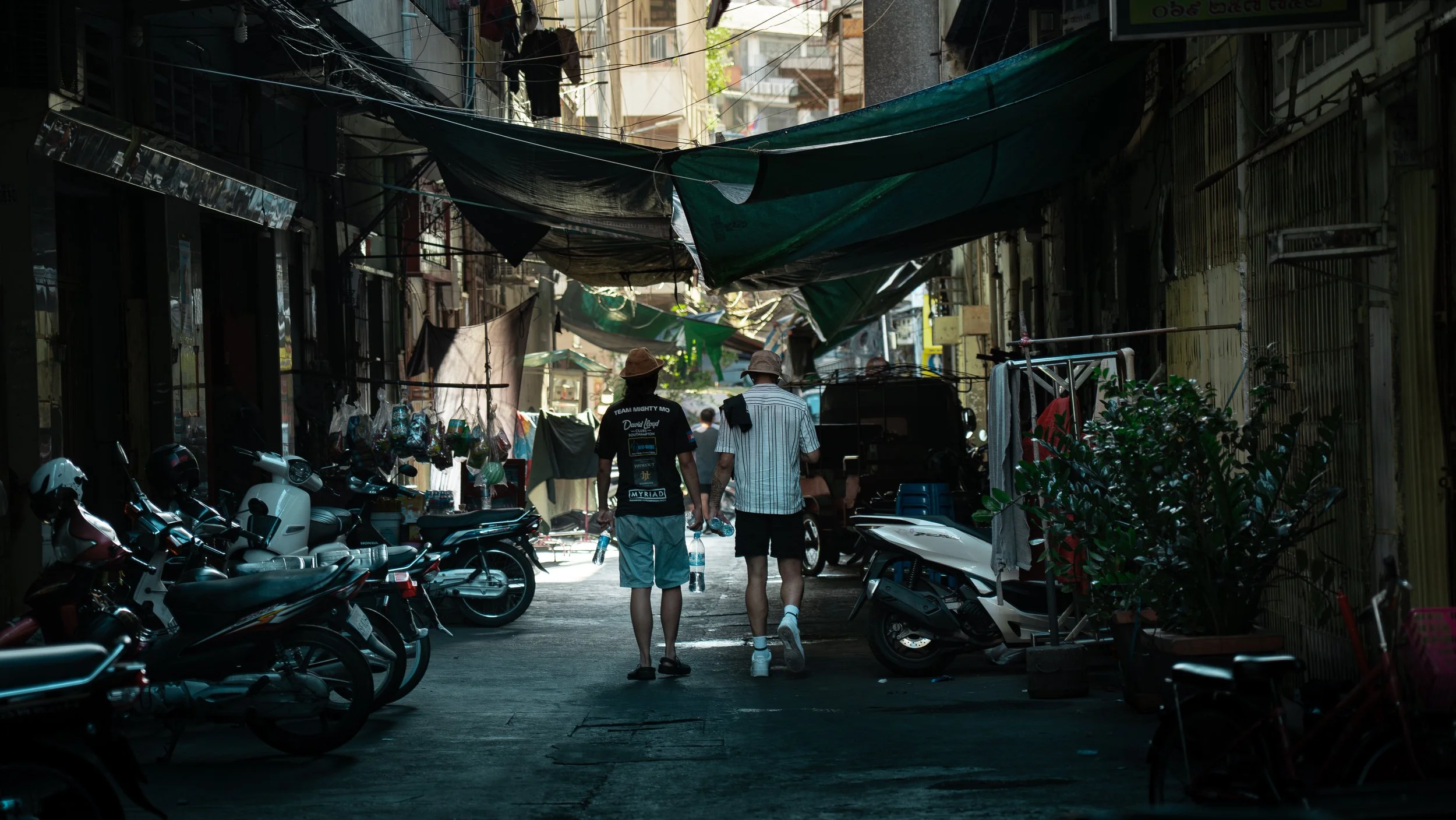 Two people walking down a narrow, shaded alleyway with motorcycles parked on both sides, small shops, hanging clothes, and makeshift canopies overhead.