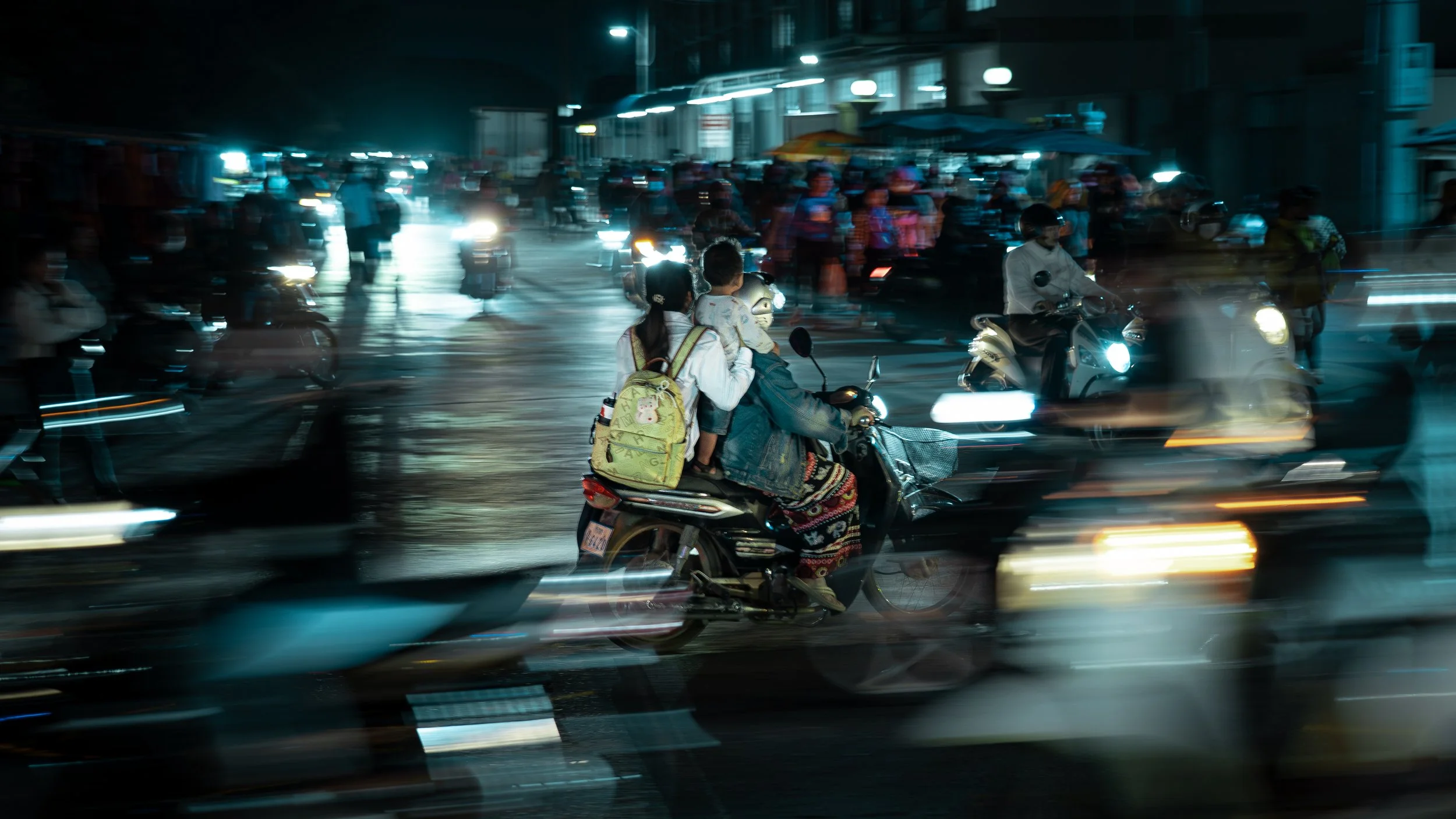 A woman riding a motorcycle with two children at night in a city, with blurred street lights and other vehicles around.