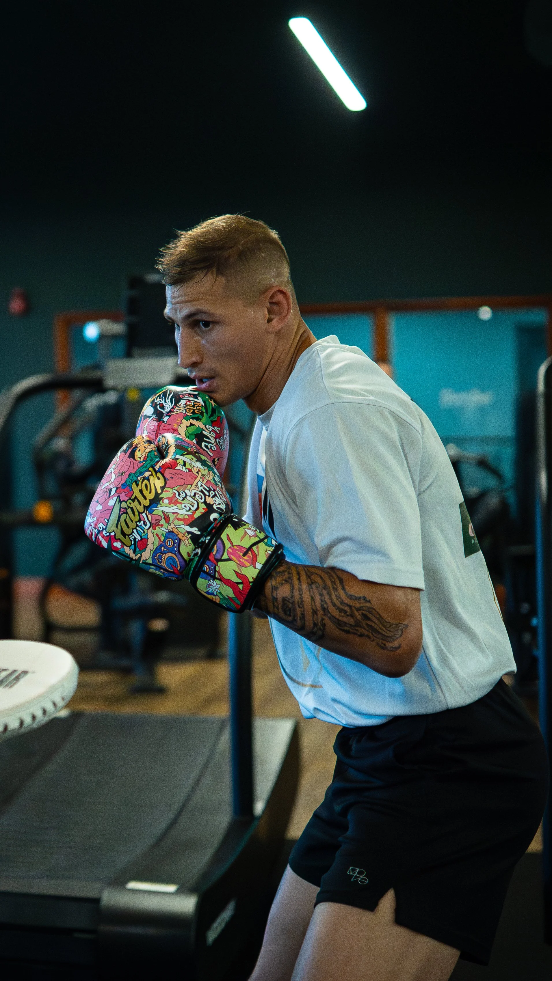 A young man with a tattoo on his arm wearing boxing gloves and sportswear in a gym, preparing to train or spar.