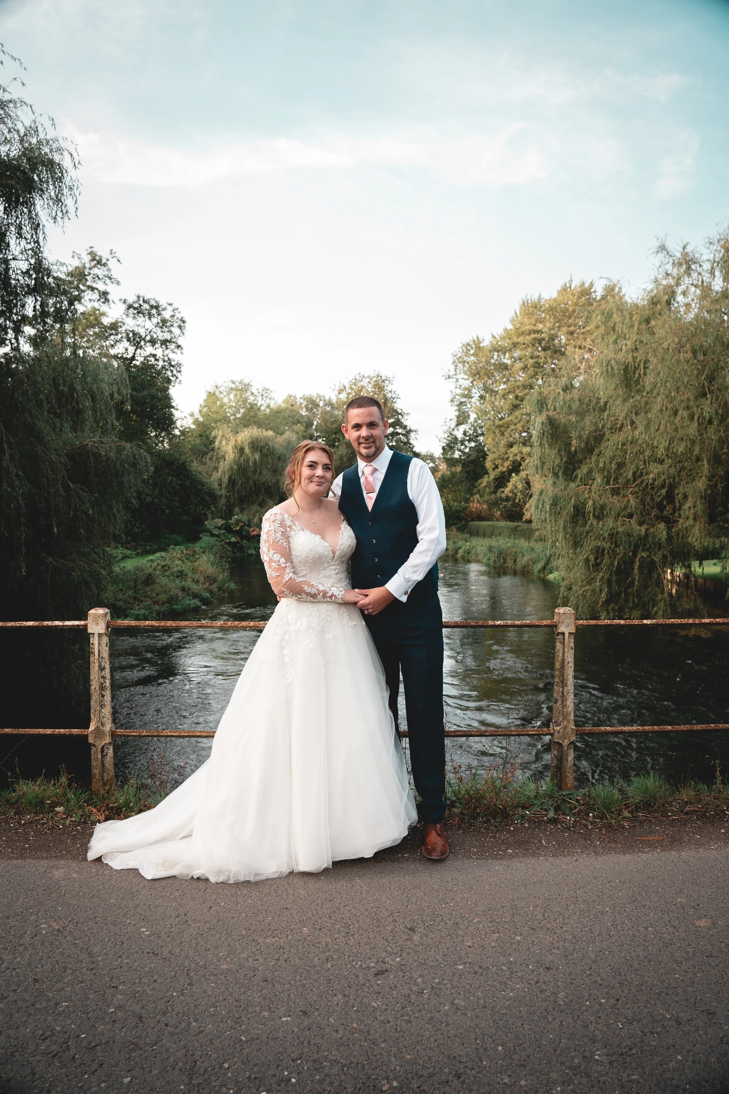 A newlywed couple standing beside a river, surrounded by lush trees, on a paved path during daytime.