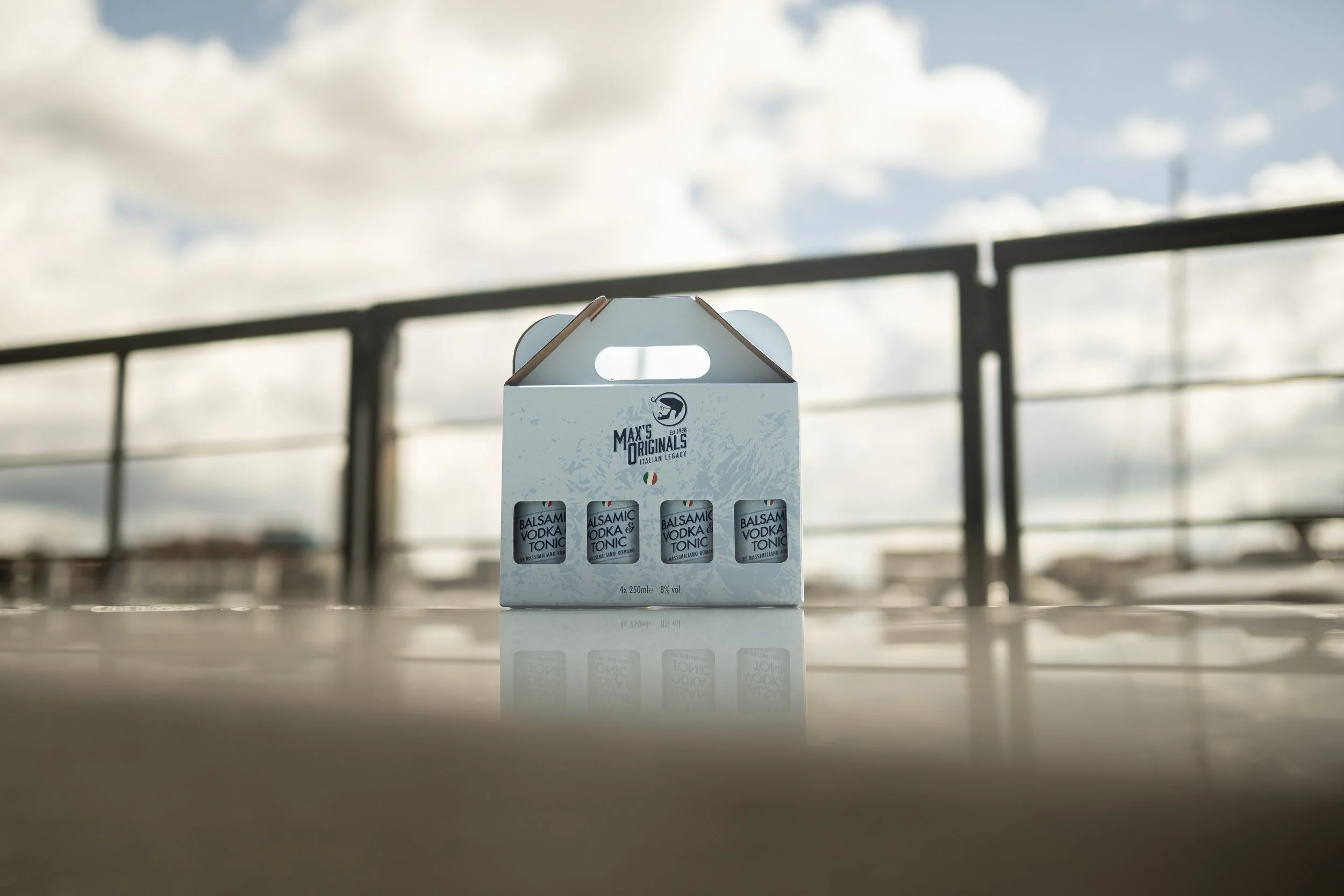 A white cardboard box containing four small bottles labeled "Balsamic Vodka & Tonic" on a reflective surface outdoors with a cloudy sky and a railing in the background.