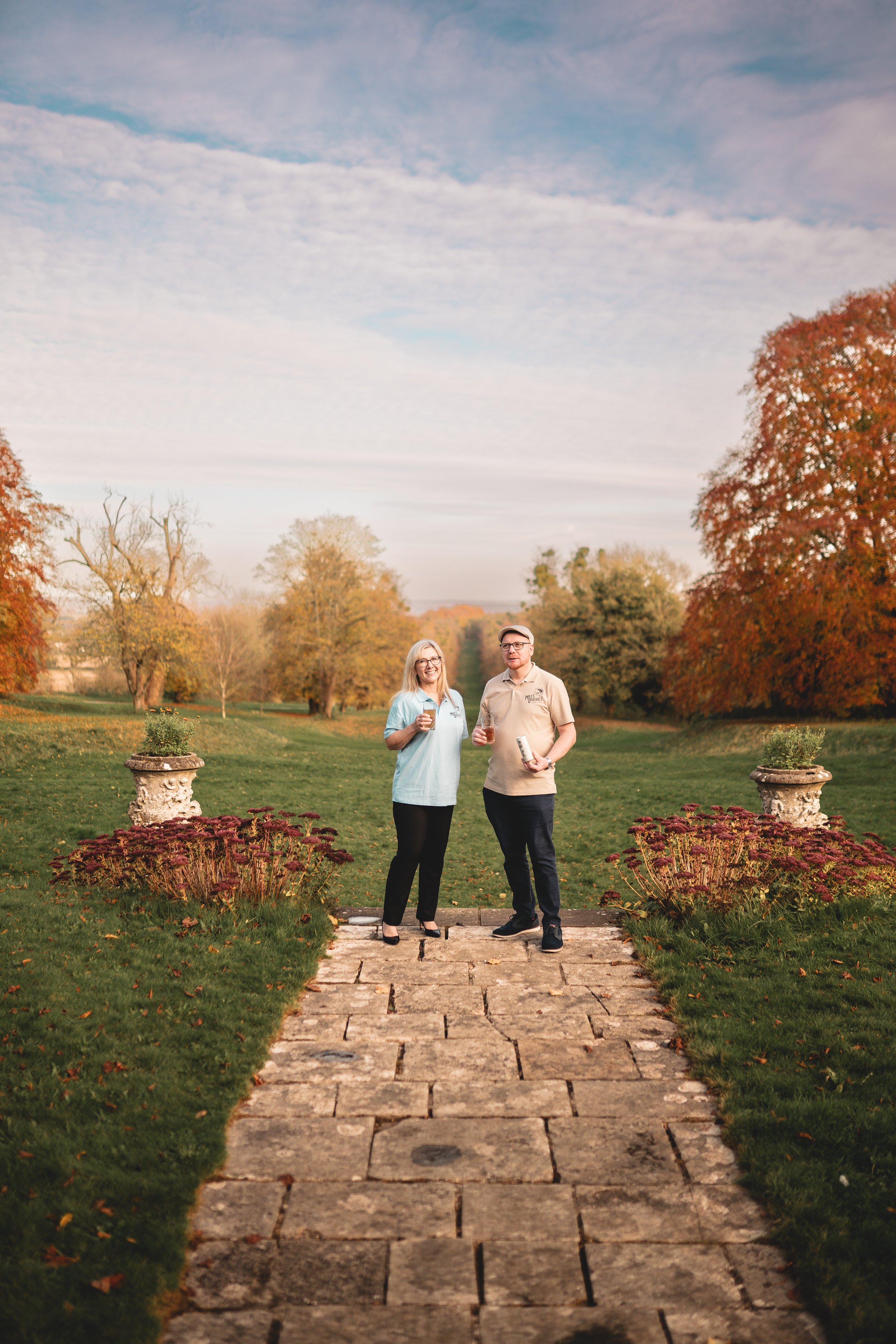 A man and a woman stand on a stone pathway in a park, holding drinks and smiling, with autumn foliage in the background.