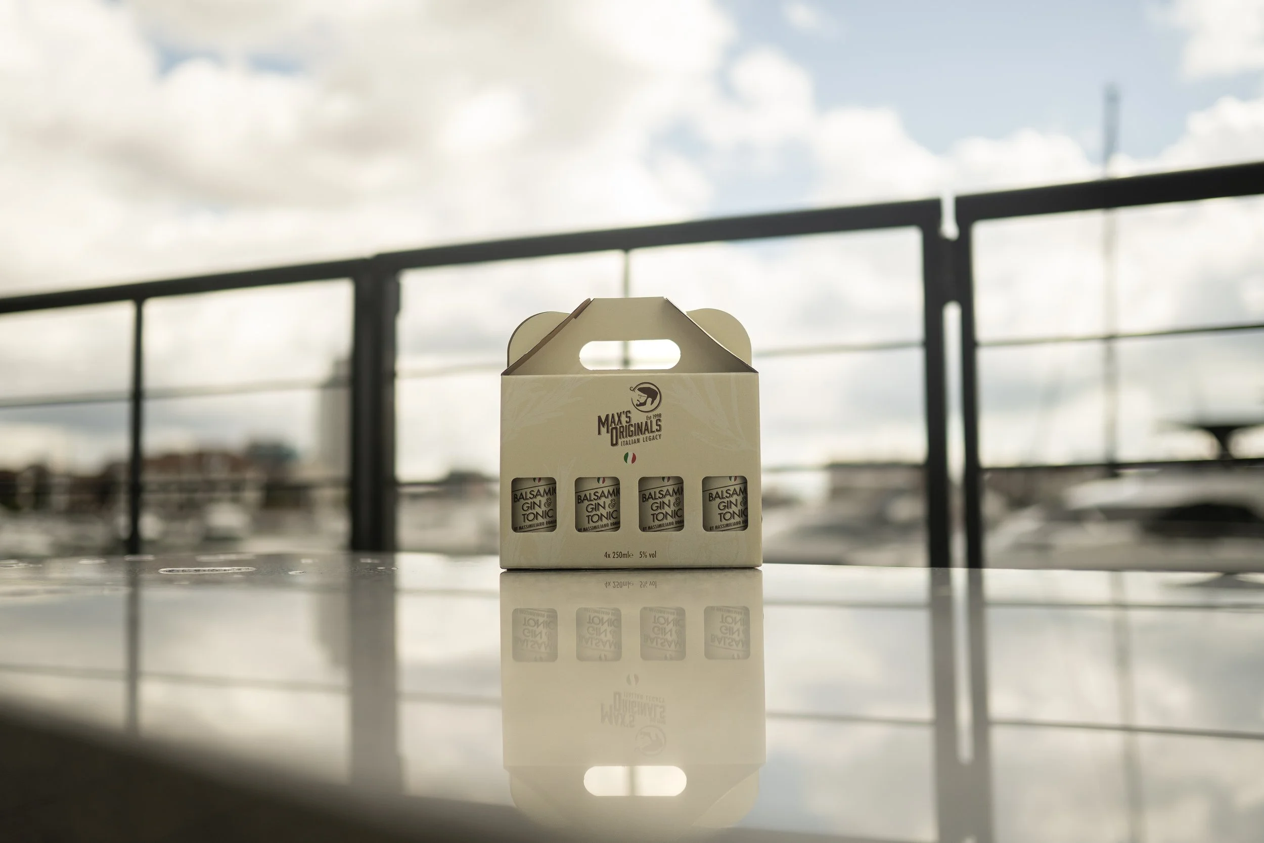 A cardboard carrier with four small bottles labeled 'Balsam GIN & TONIC' placed on a reflective surface outdoors with a blurred skyline and clouds in the background.
