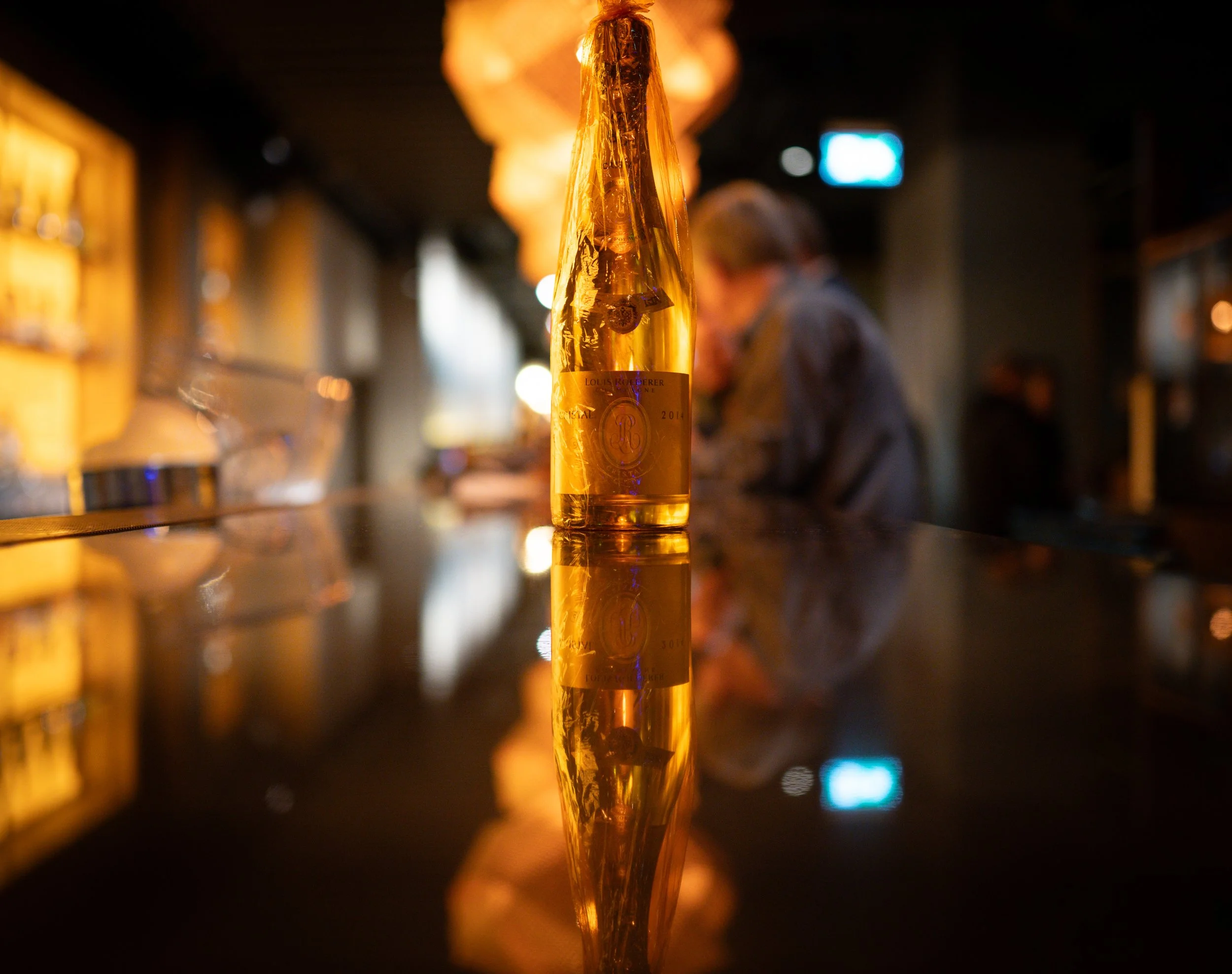A yellow wine bottle with a gold label on a reflective bar counter in a dimly lit bar or restaurant with warm lighting and blurred background of people and shelves.