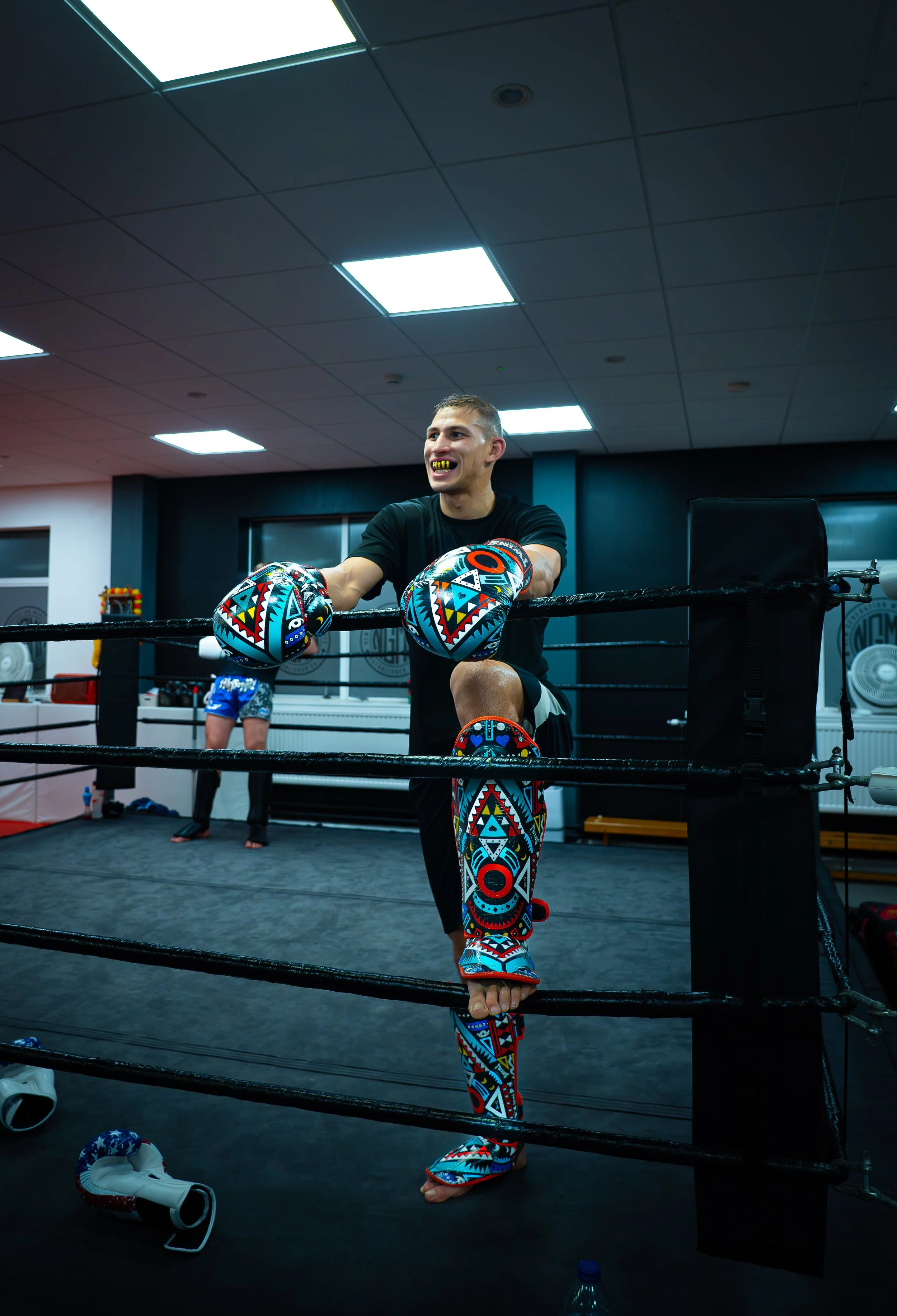 A young man training in a boxing gym, wearing colorful patterned boxing gloves, shin guards, and mouthguard, leaning on the ropes of a boxing ring, with another person in the background and gym equipment around.