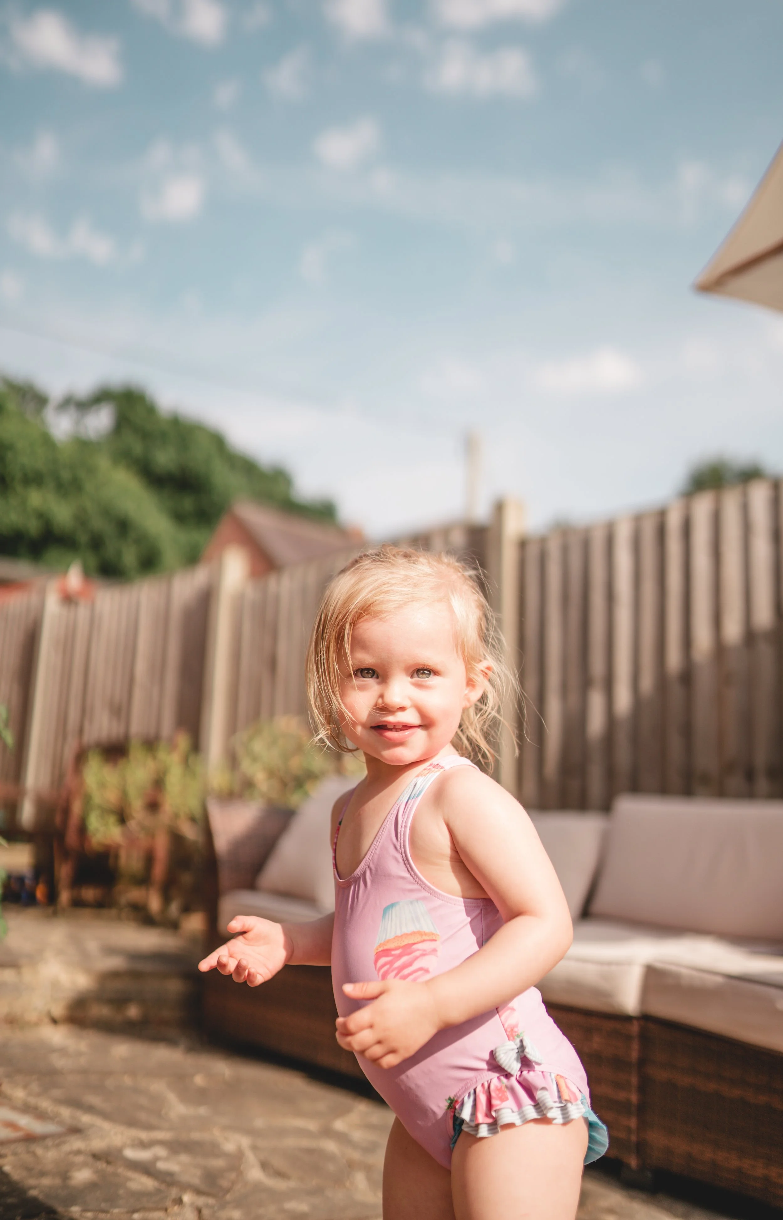 A young girl with blonde hair and blue eyes smiling outdoors in a backyard on a sunny day, wearing a pink swimsuit with ice cream cone graphics and ruffle details.