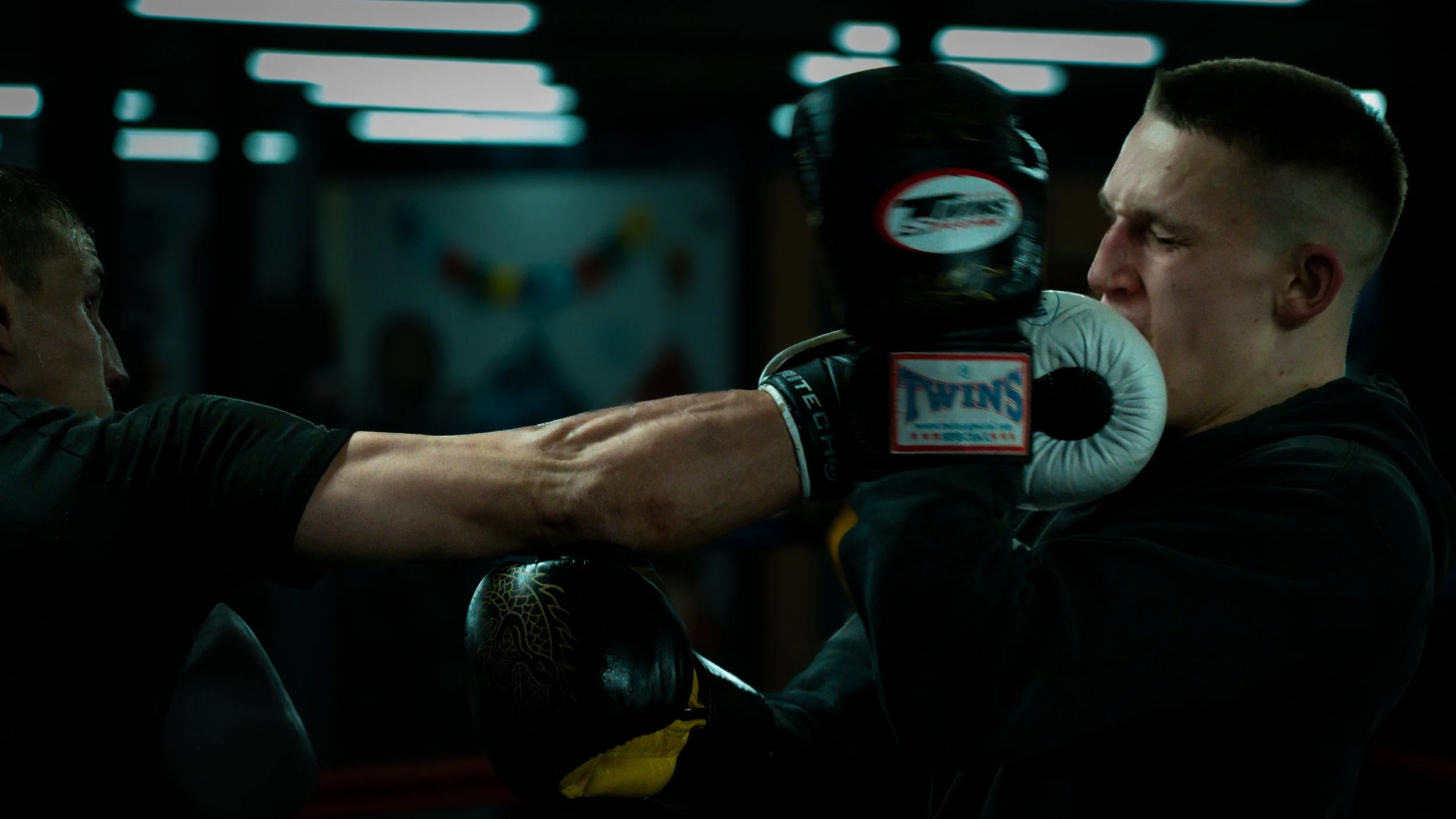 Two men training in a boxing gym, one man is throwing a punch towards the other, who is wearing boxing gloves and holding up a padded target for practice.