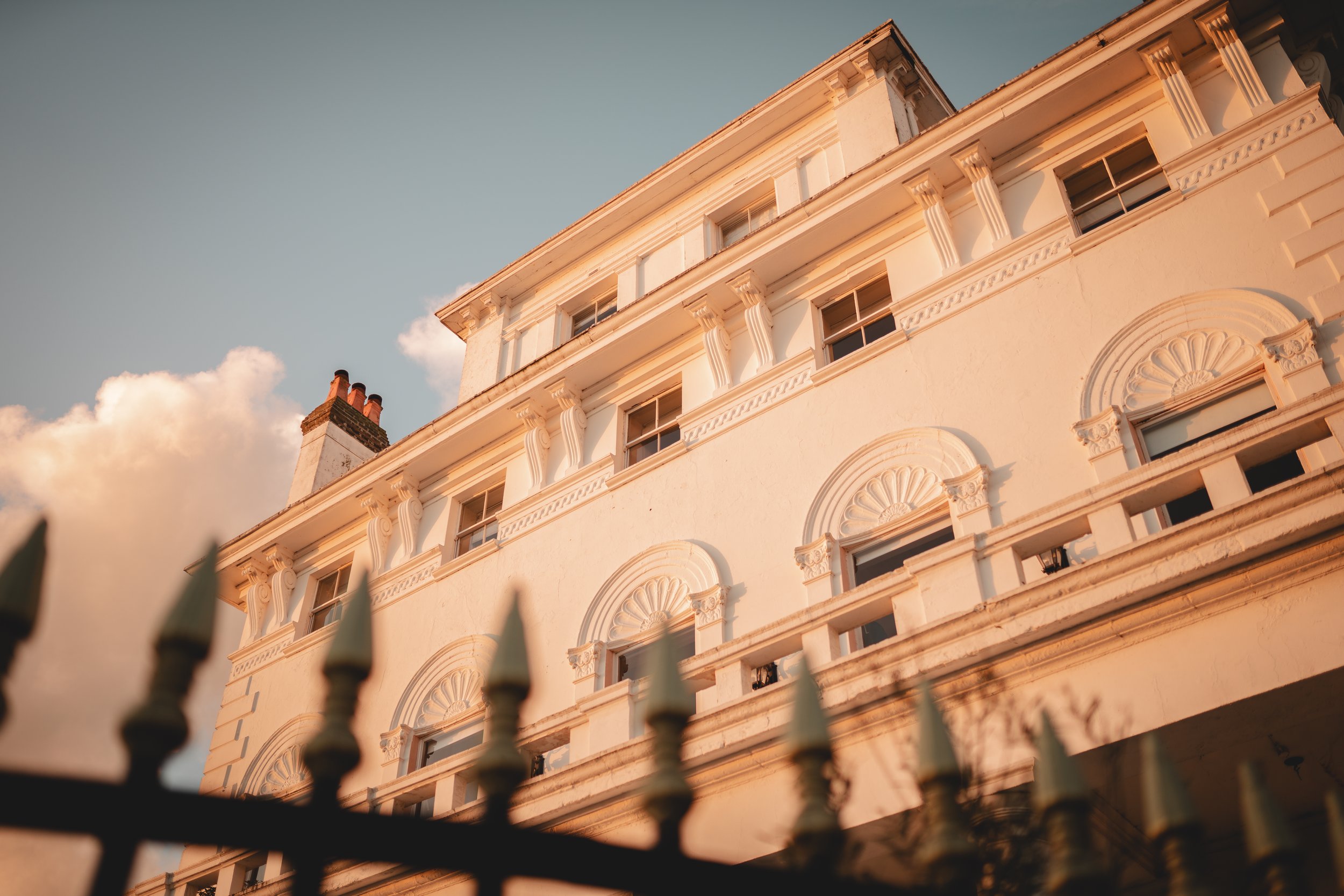Low-angle view of a white historic building with classical architecture details, sunlight illuminating its facade, iron fence in foreground, partly cloudy sky in the background.