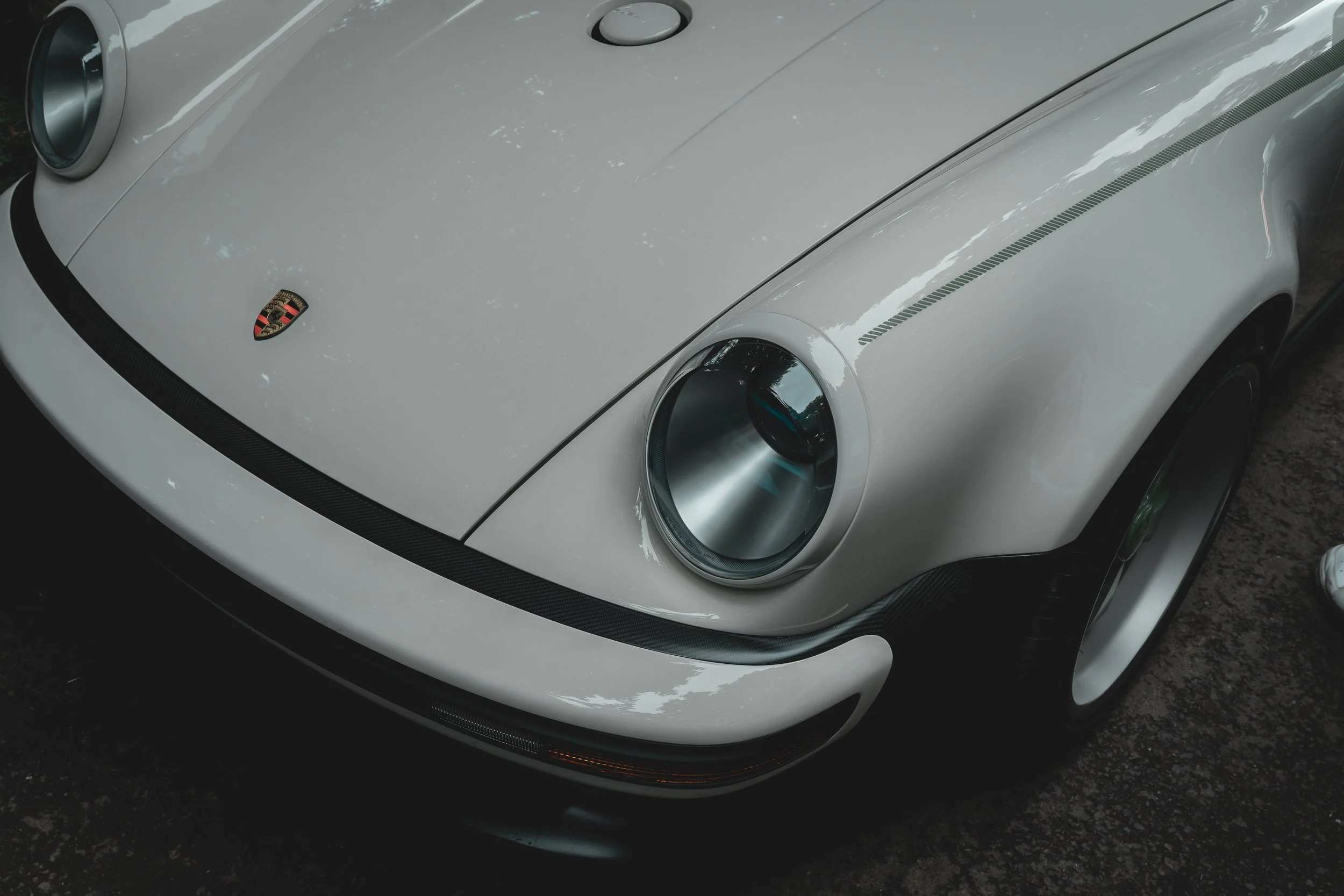 Close-up of a white vintage sports car, focusing on the front view. The car features round headlights, a Porsche badge on the hood, and black trim details.