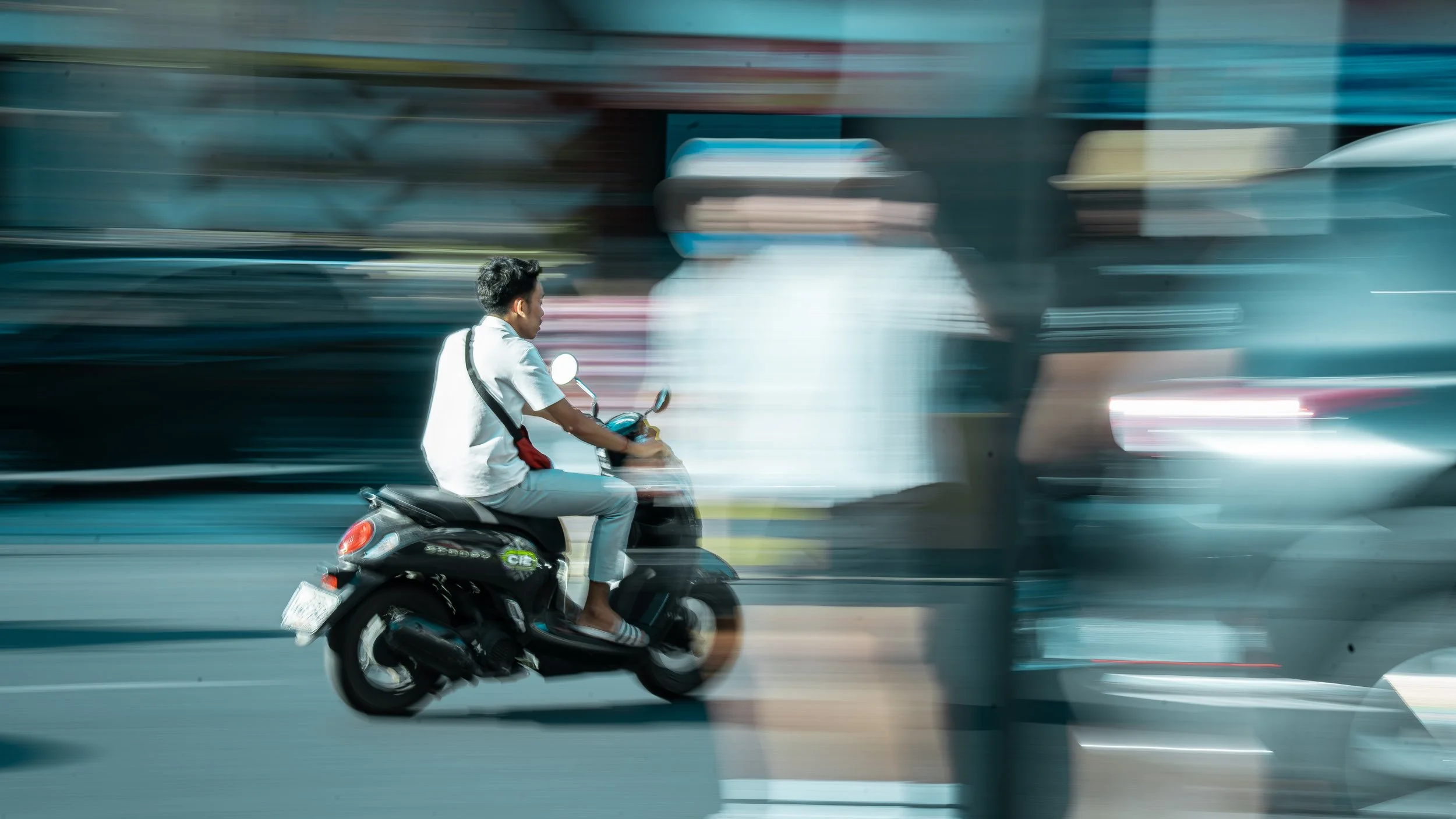 A man riding a black scooter on a city street with blurred vehicles and buildings in the background.