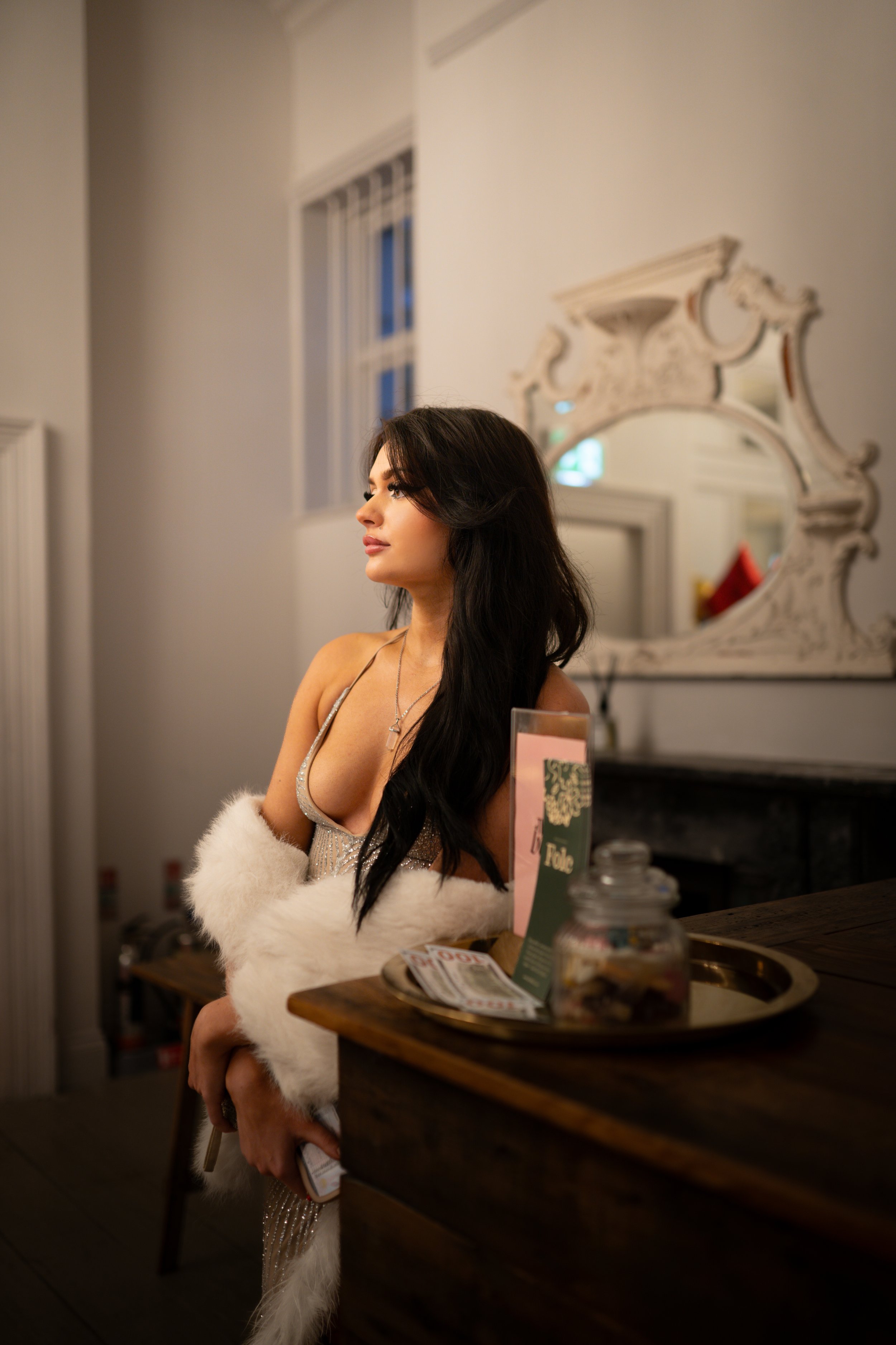 A woman in a glamorous silver dress and fur stole sitting at a wooden table in a warmly lit room with ornate decor, a mirror on the wall, and a window in the background.