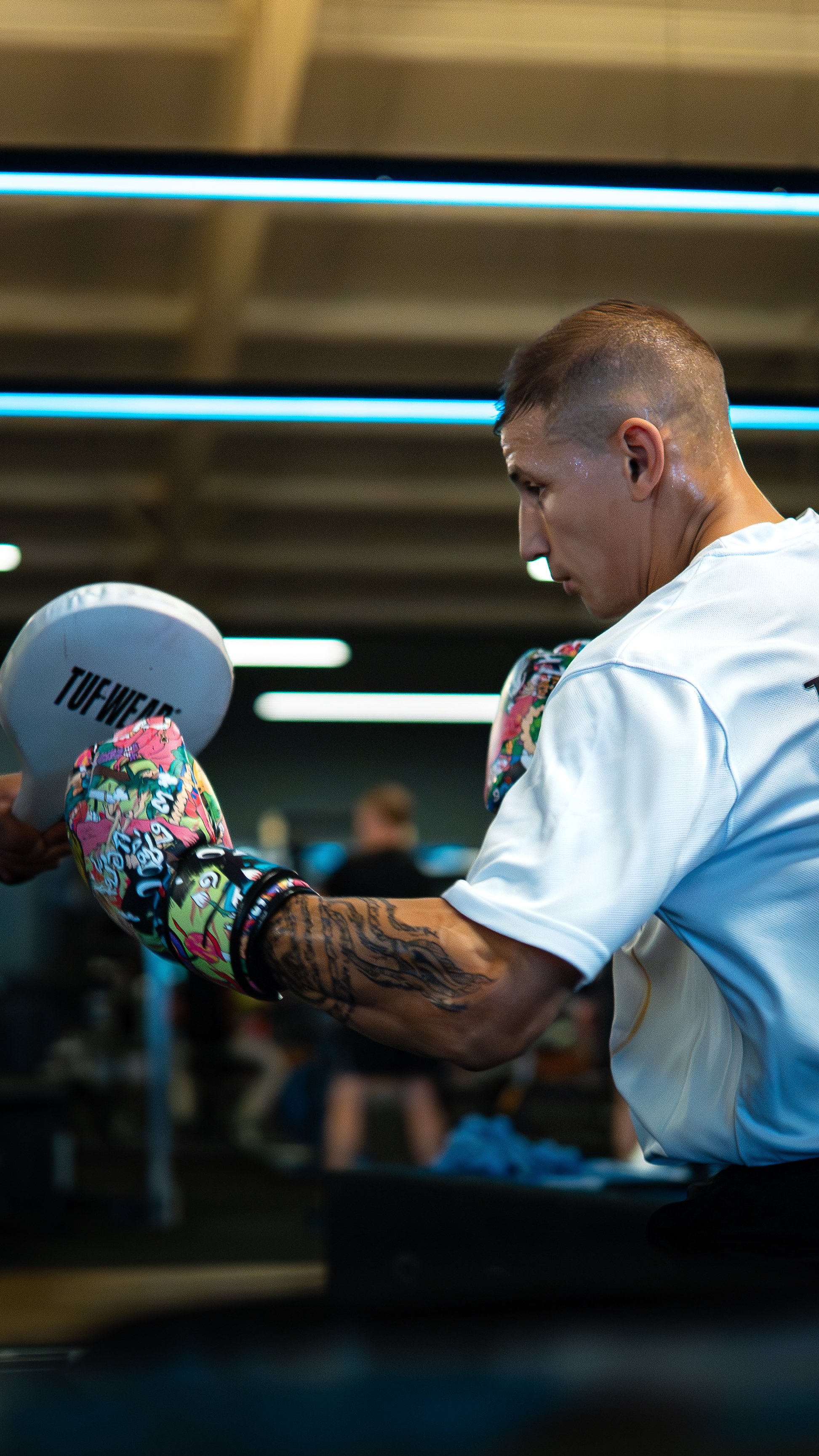 A male boxer wearing colorful boxing gloves, white shirt, and with tattoos on his arm, practicing his punch in a gym.