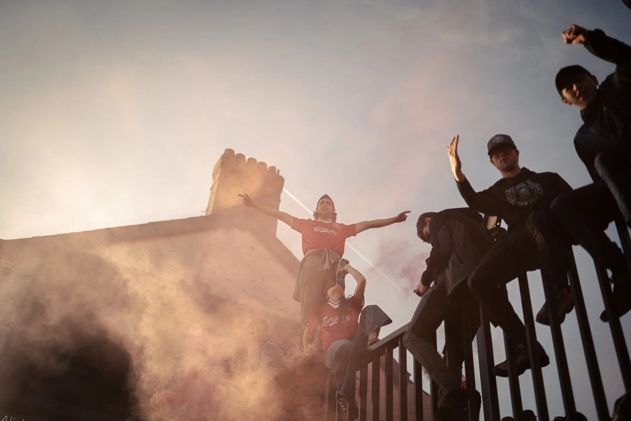 Group of young men on a balcony with smoke or fog, one with arms outstretched, some raising their hands, against a cloudy sky.
