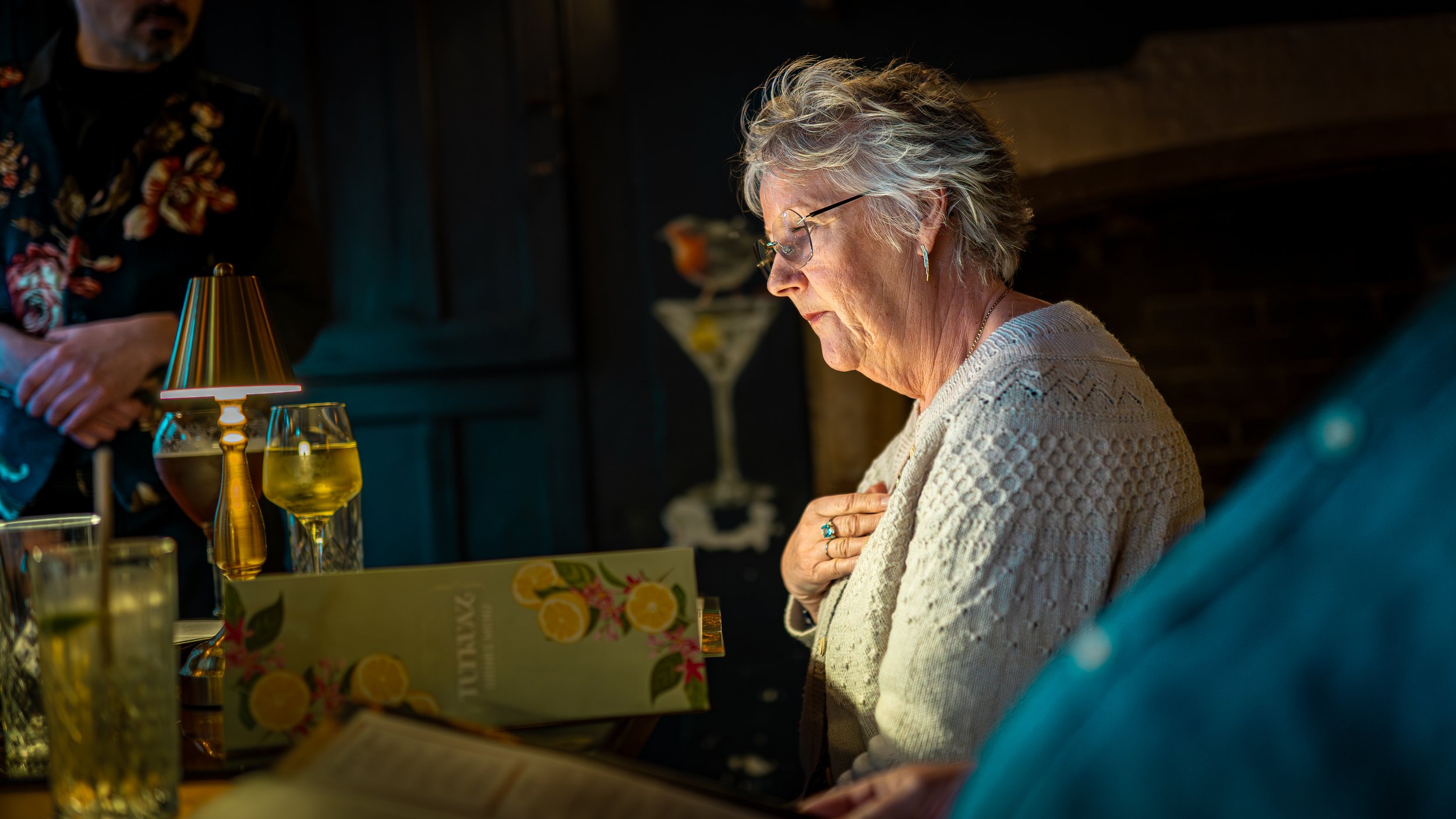 An elderly woman with glasses and short gray hair, dressed in a light-colored knitted sweater, sits at a table with drinks and a menu, looking down with a thoughtful expression, in a dimly lit room.