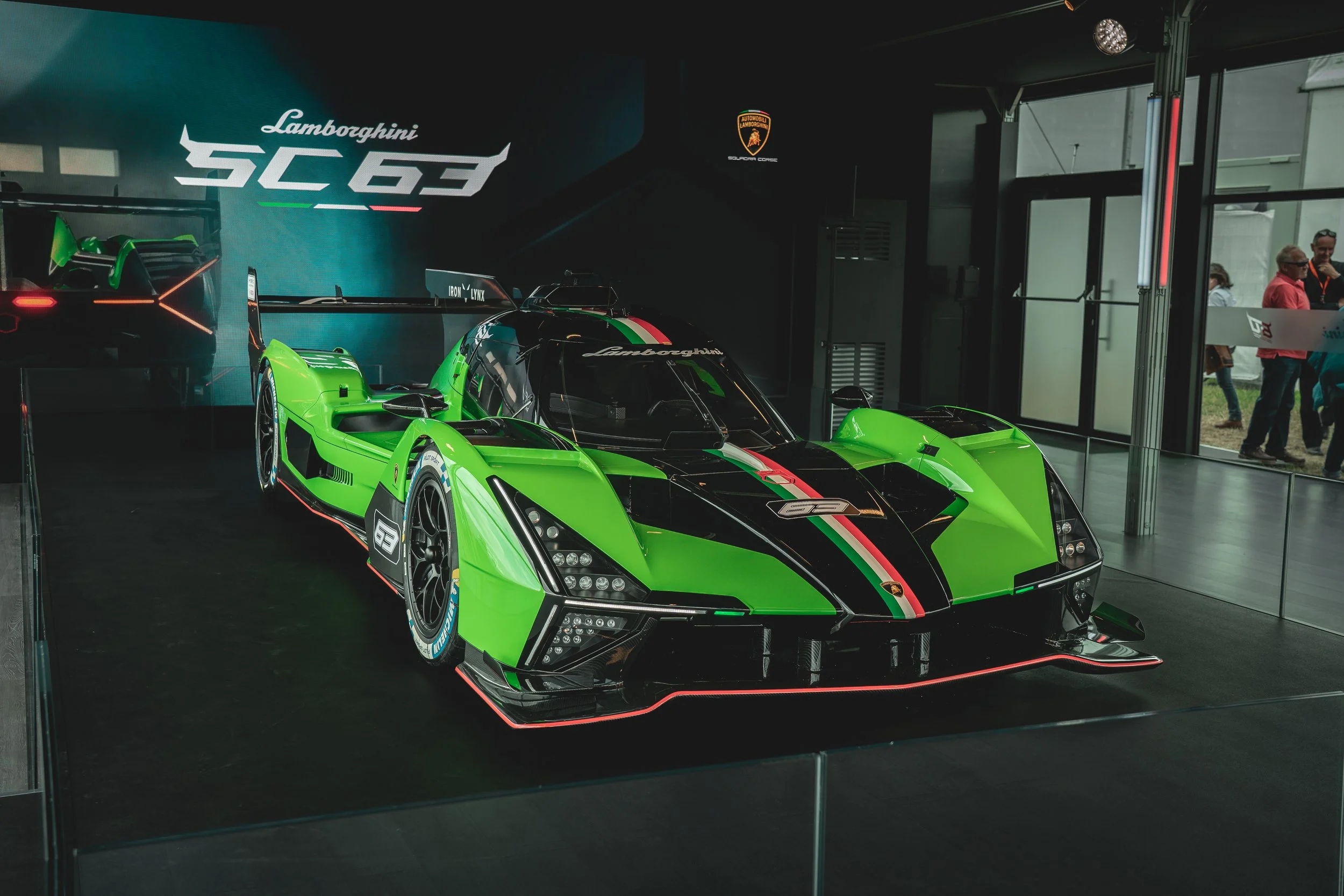 Green and black Lamborghini SC63 race car displayed indoors, with 'Lamborghini' logo and 'SC63' on background, surrounded by visitors.