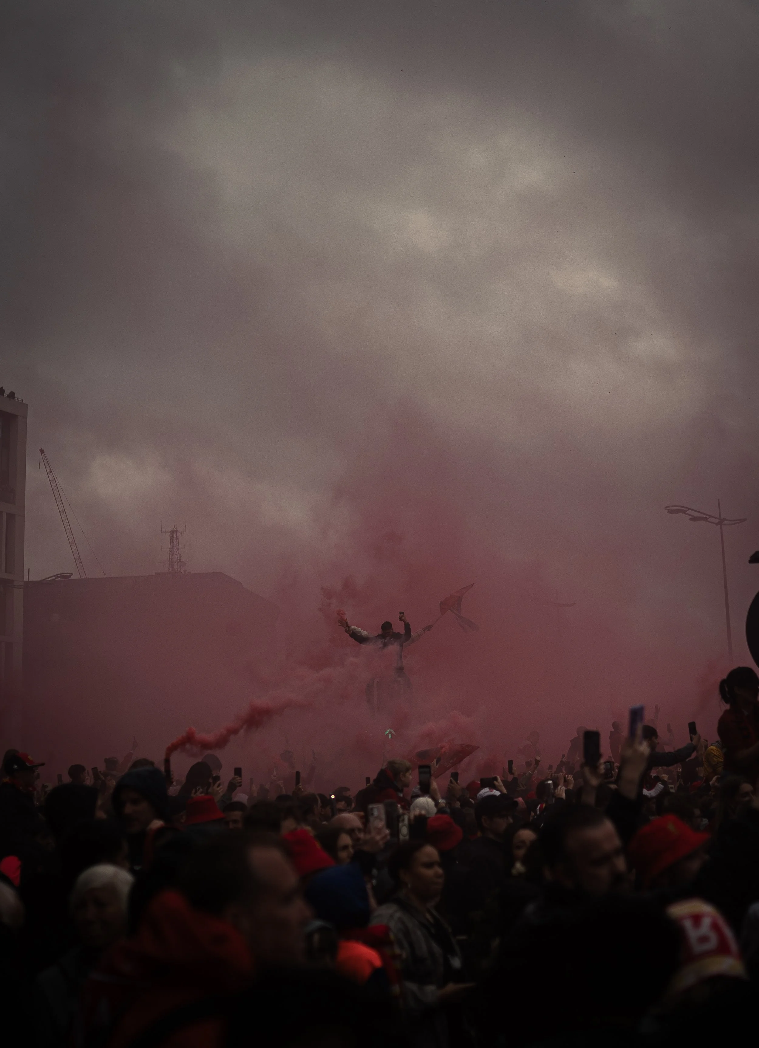 Crowd of people gathered outdoors with a man on a bike holding a flag, emitting pink smoke, during a protest or celebration, with a cloudy sky overhead.