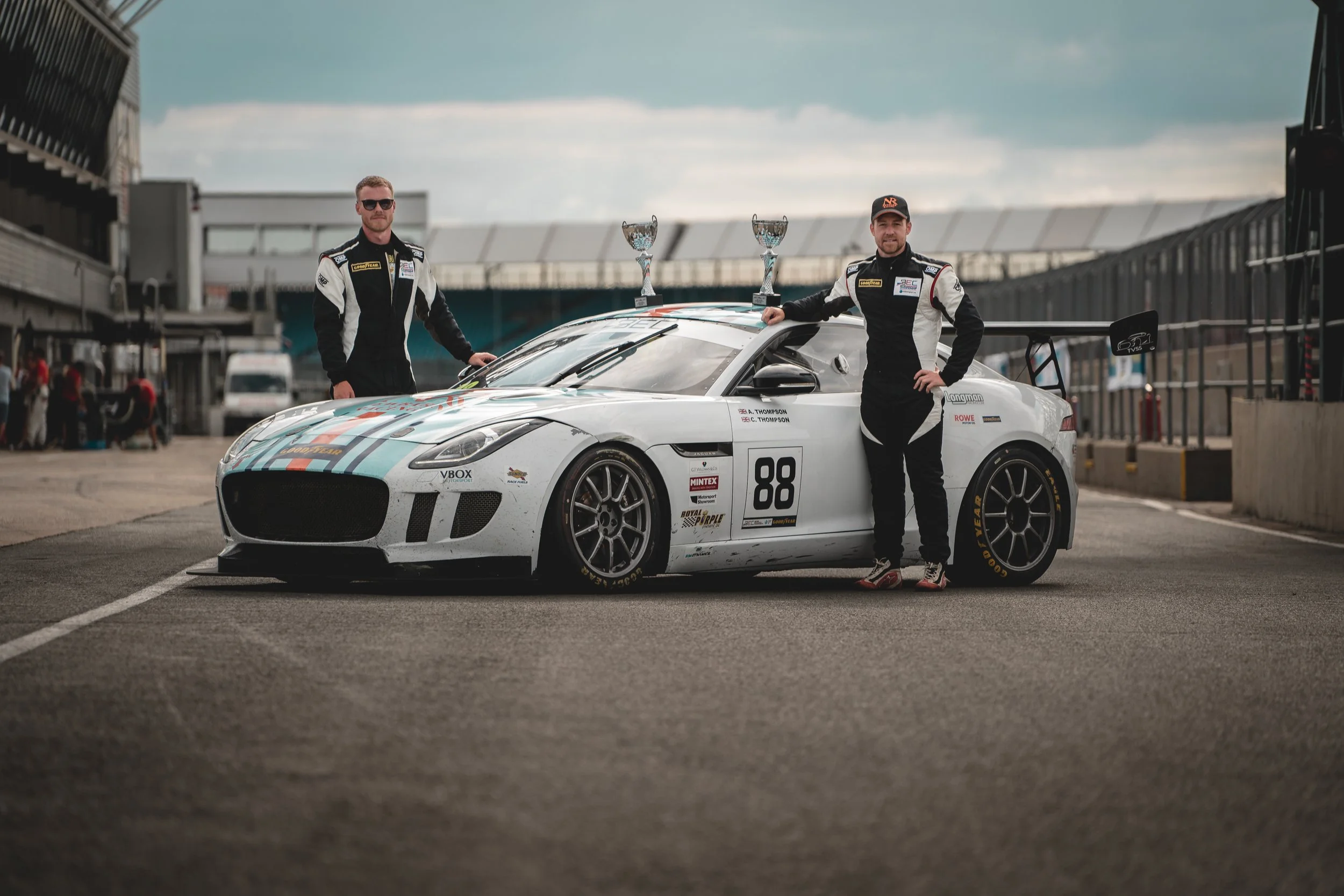 Two race car drivers standing next to a white racing car with the number 88, holding trophies, in a race track pit area.
