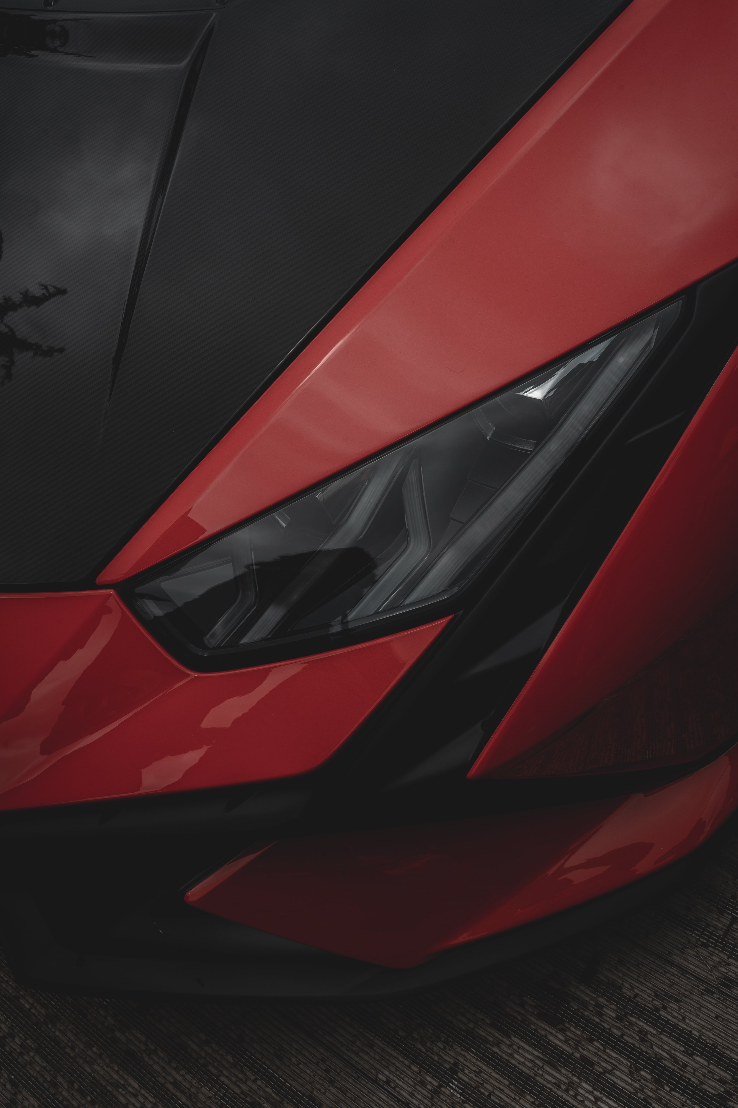 Close-up of the front part of a red sports car with a black carbon fiber hood and sleek headlight design.