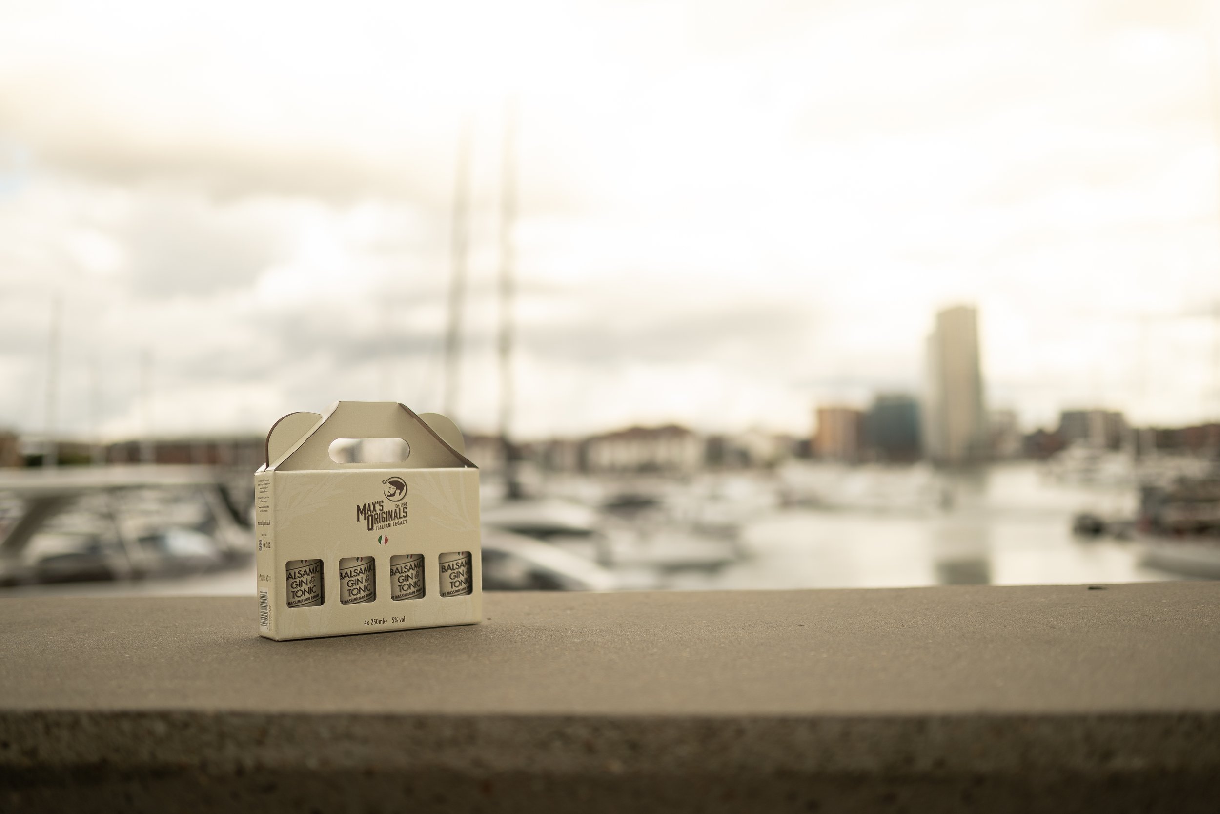 Packaged bottles of balsamic gin tonic on a concrete surface with a blurred marina and cityscape in the background.