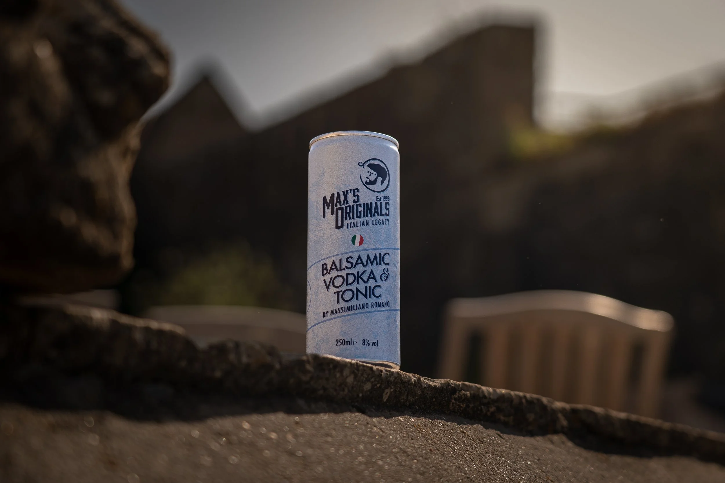 A can of Max's Originals balsamic vodka and tonic drink standing on a ledge outdoors with a blurred background of rocks and furniture.