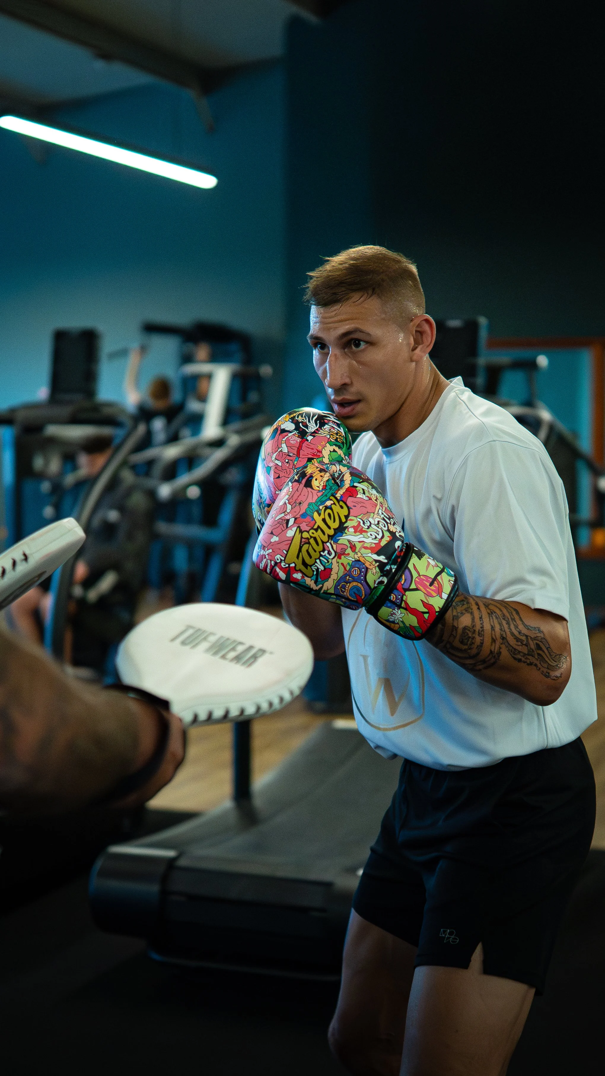 A man training with boxing gloves and mitts in a gym, with weight machines in the background.