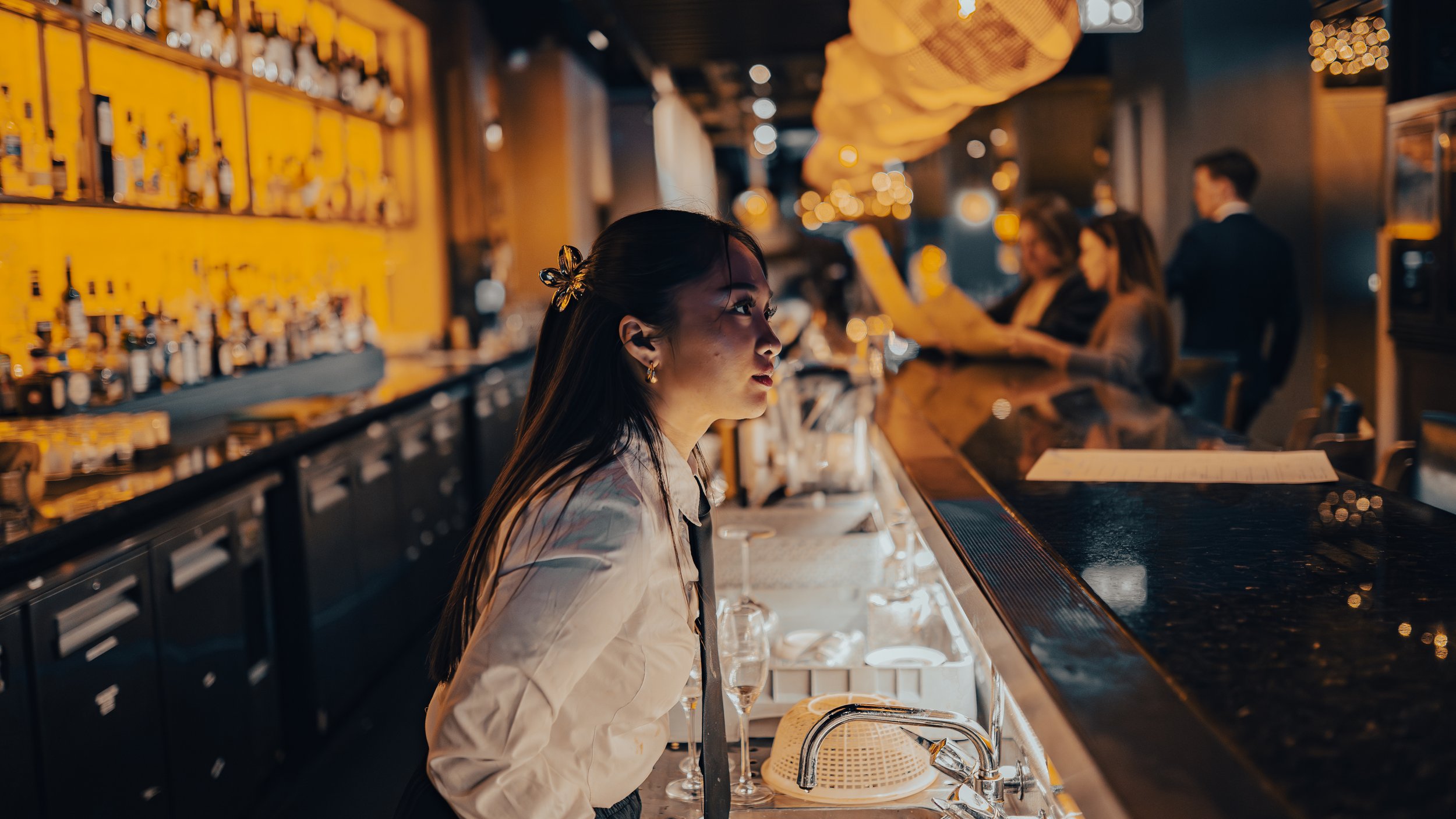 A woman bartending at a bar, lit with warm lighting, with bottles displayed on yellow shelves behind her. Other patrons are sitting at the bar, some reading menus or chatting.