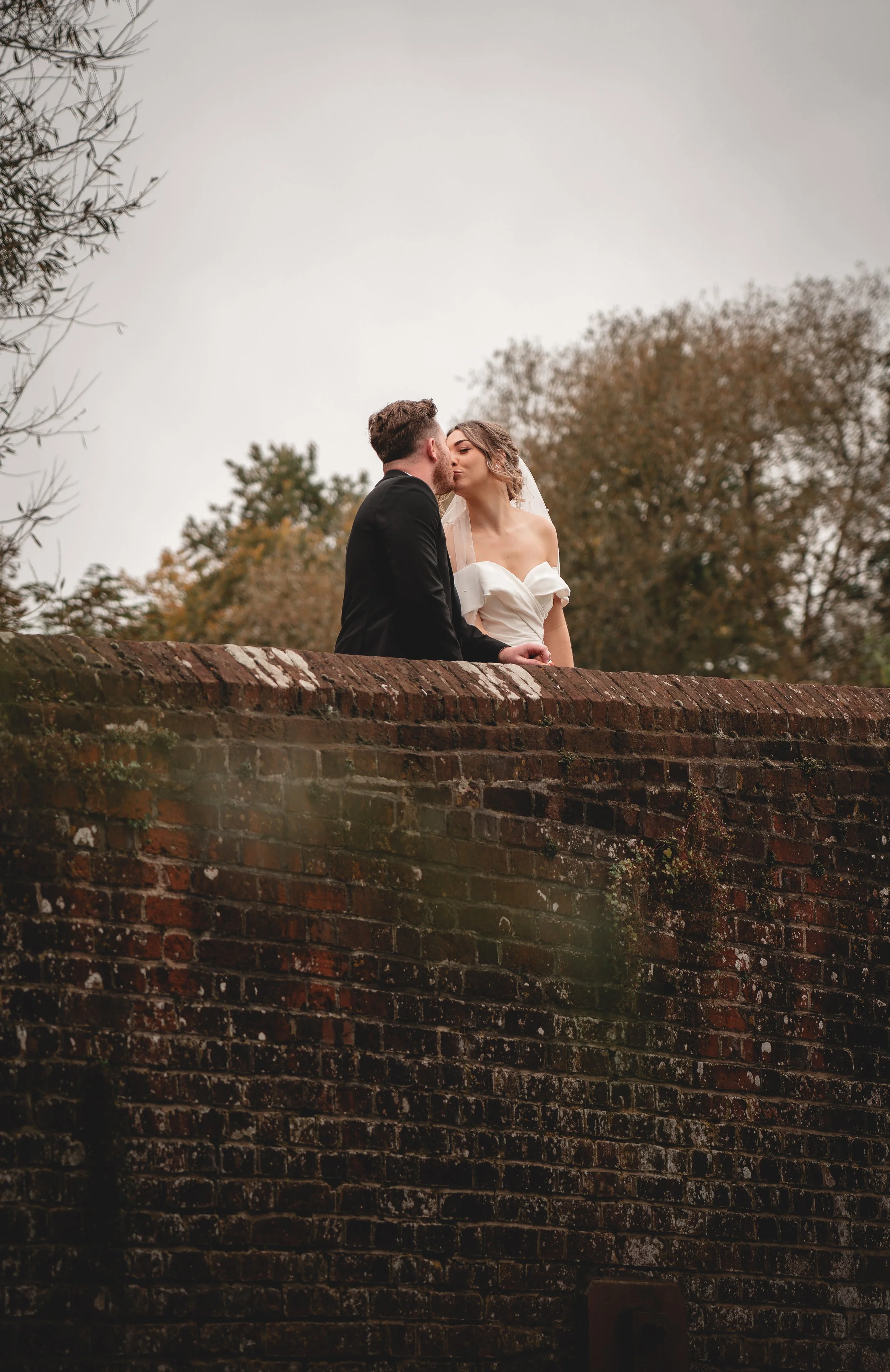 A bride and groom share a kiss on a brick bridge surrounded by trees, during an outdoor wedding photoshoot.