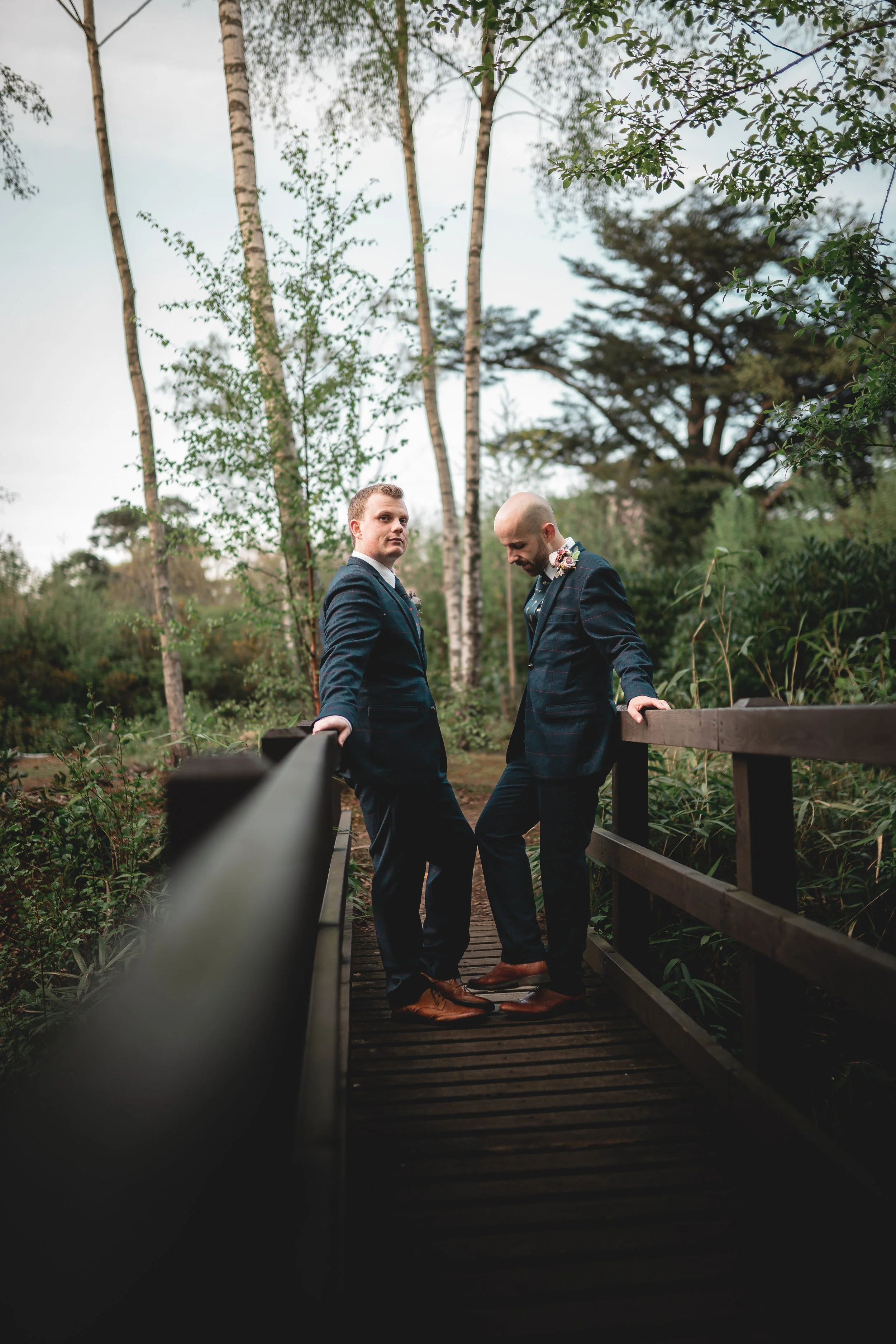 Two men in suits standing on a wooden bridge in a forested area, looking at the camera.