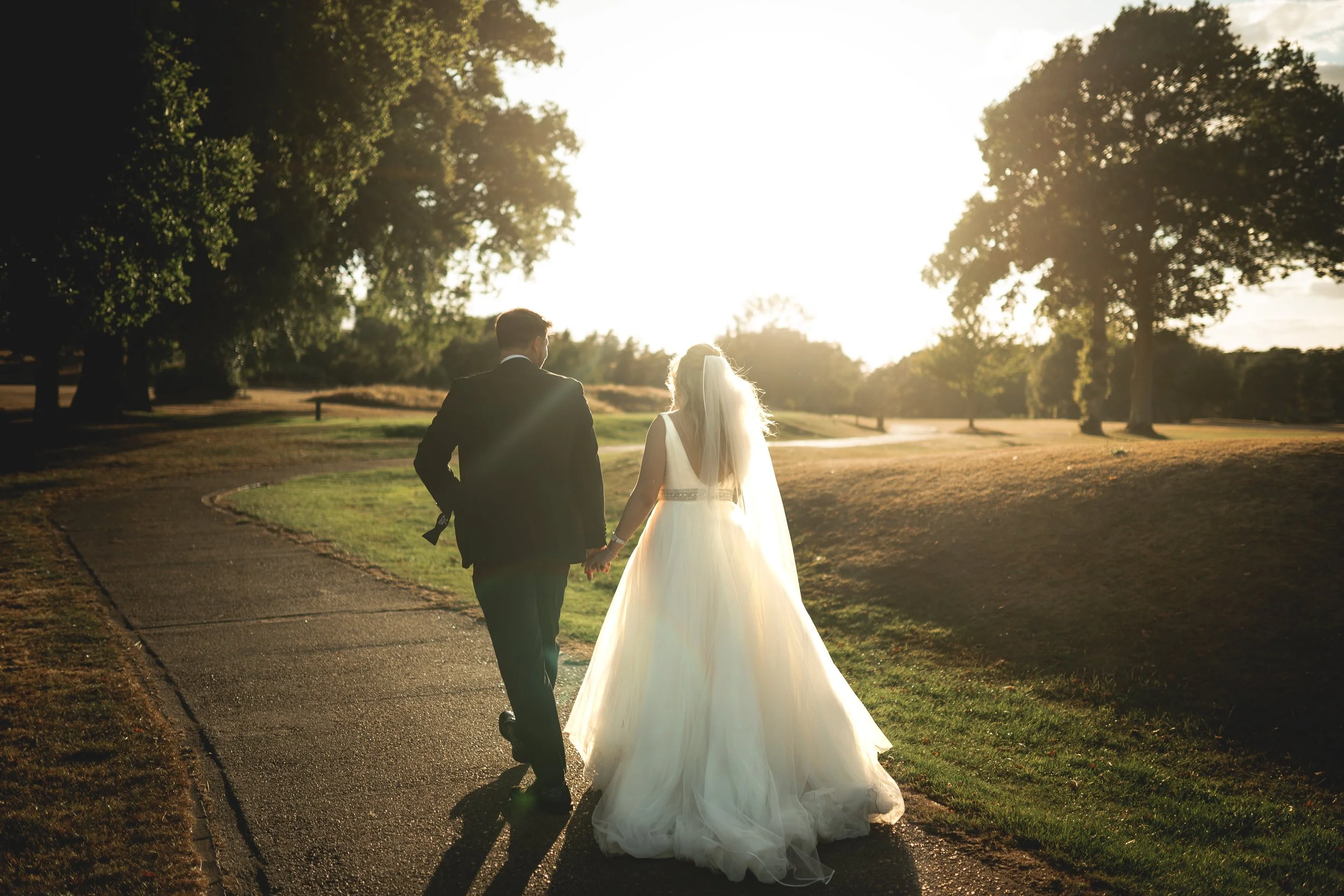 A bride and groom walking hand in hand on a paved path in a park during sunset.