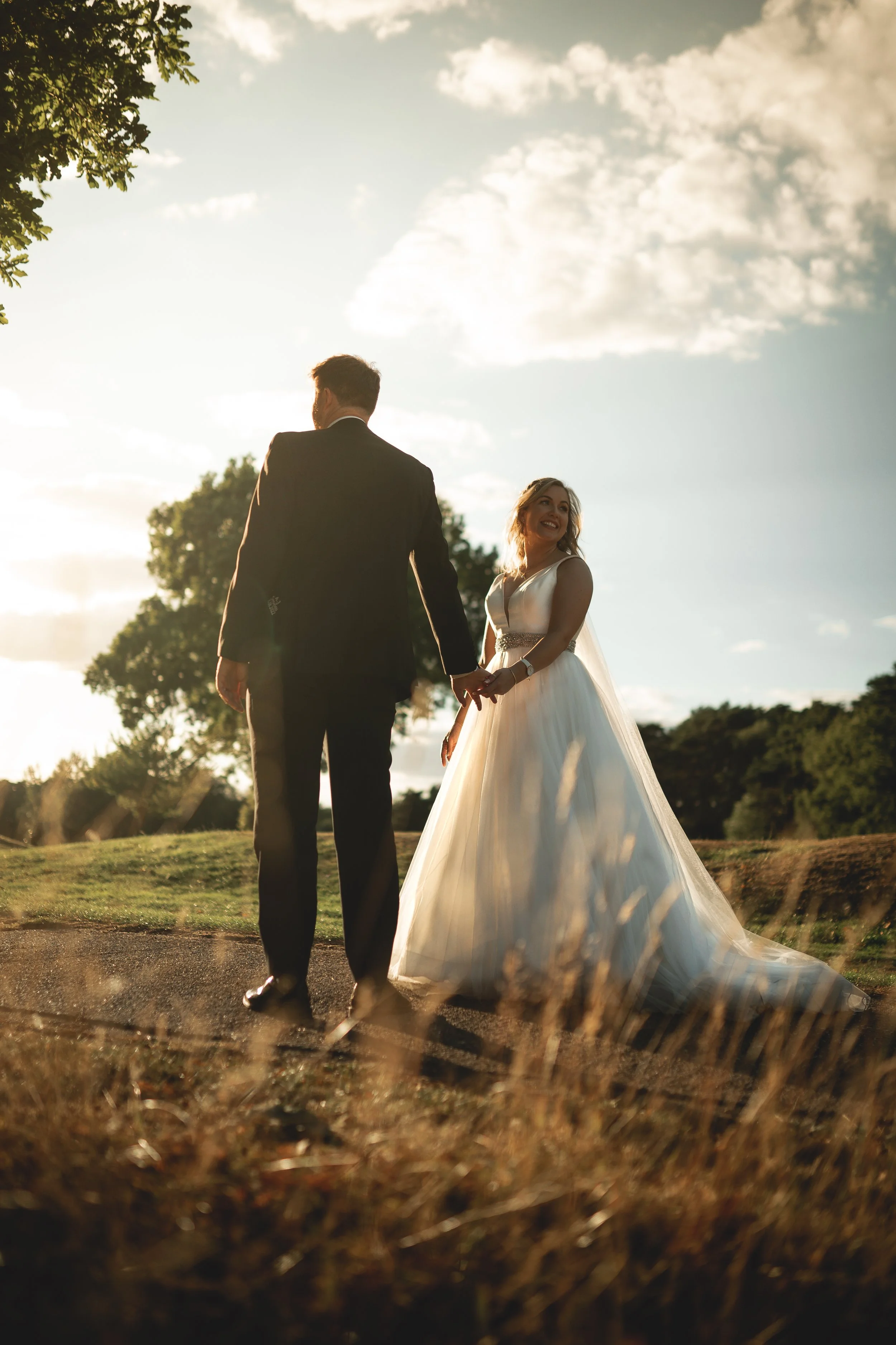 A bride and groom holding hands outdoors during sunset, with the bride smiling and wearing a white wedding dress, and the groom in a black suit.