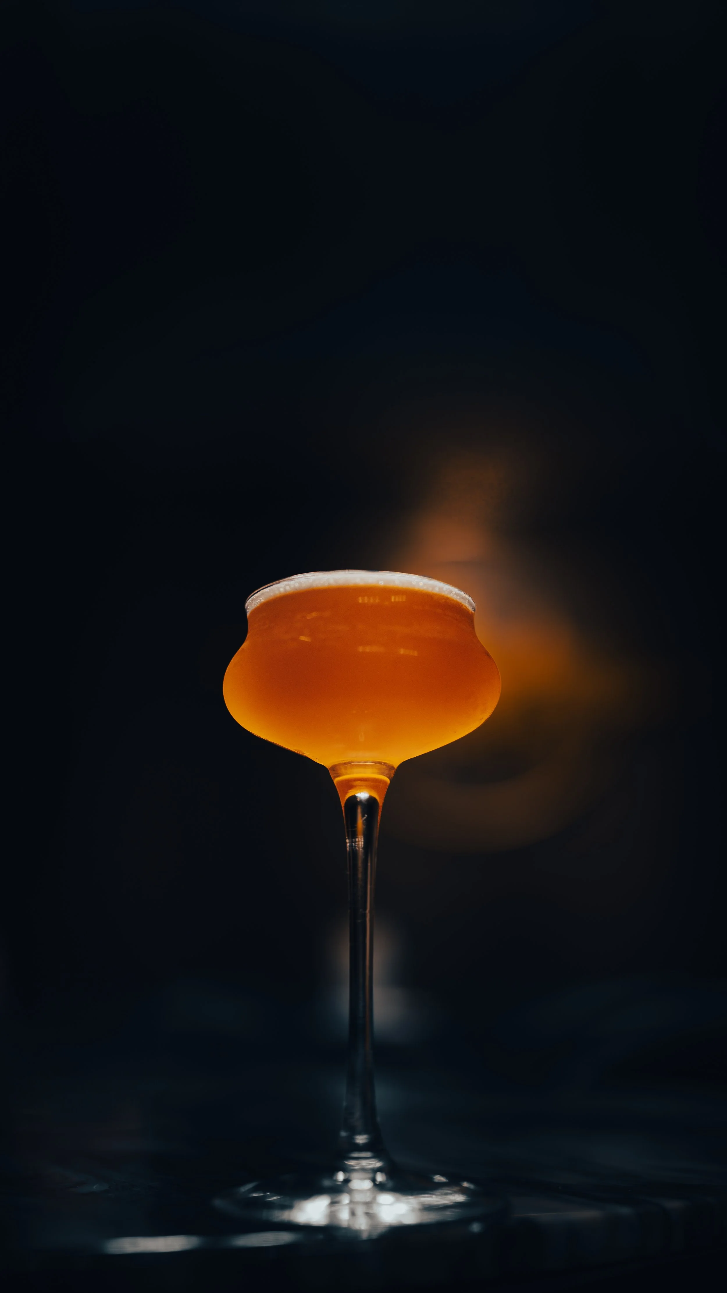 A cocktail glass filled with an amber-colored drink, placed against a dark background.