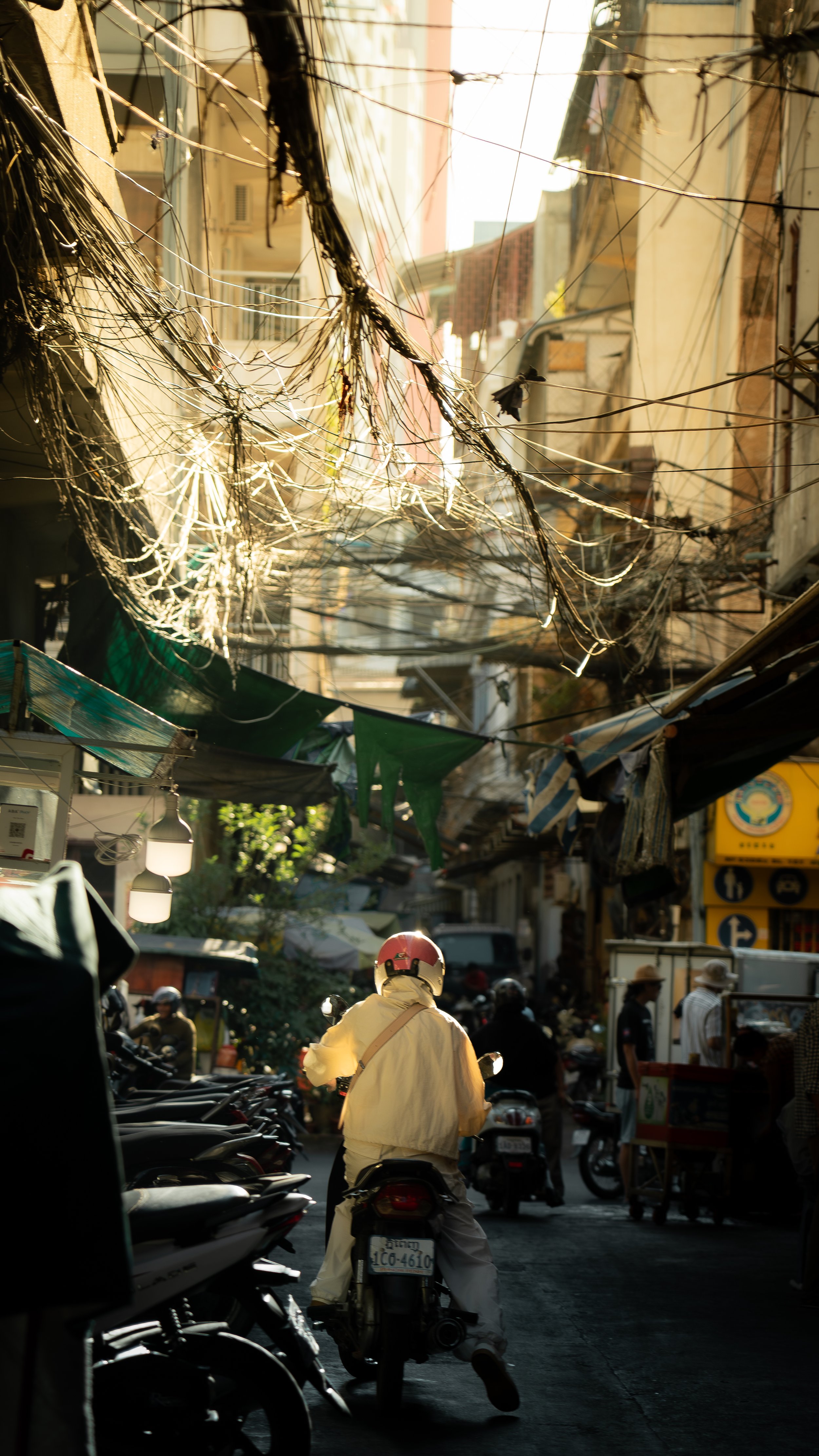 A busy street scene in a Southeast Asian city, with a person on a scooter in the foreground and tangled electrical wires overhead, storefronts, and people walking in the background.