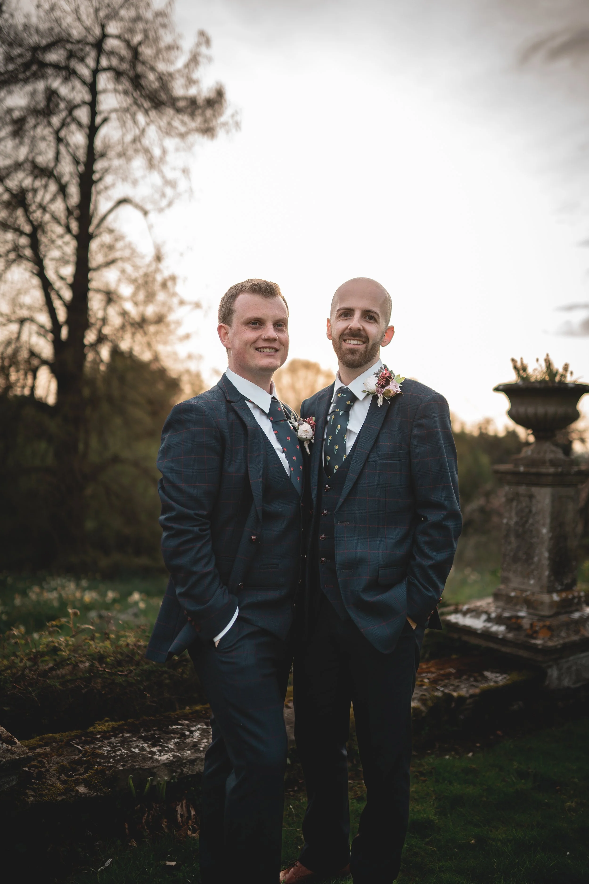 Two men dressed in formal suits standing outdoors at sunset, smiling, with trees and a stone column in the background.