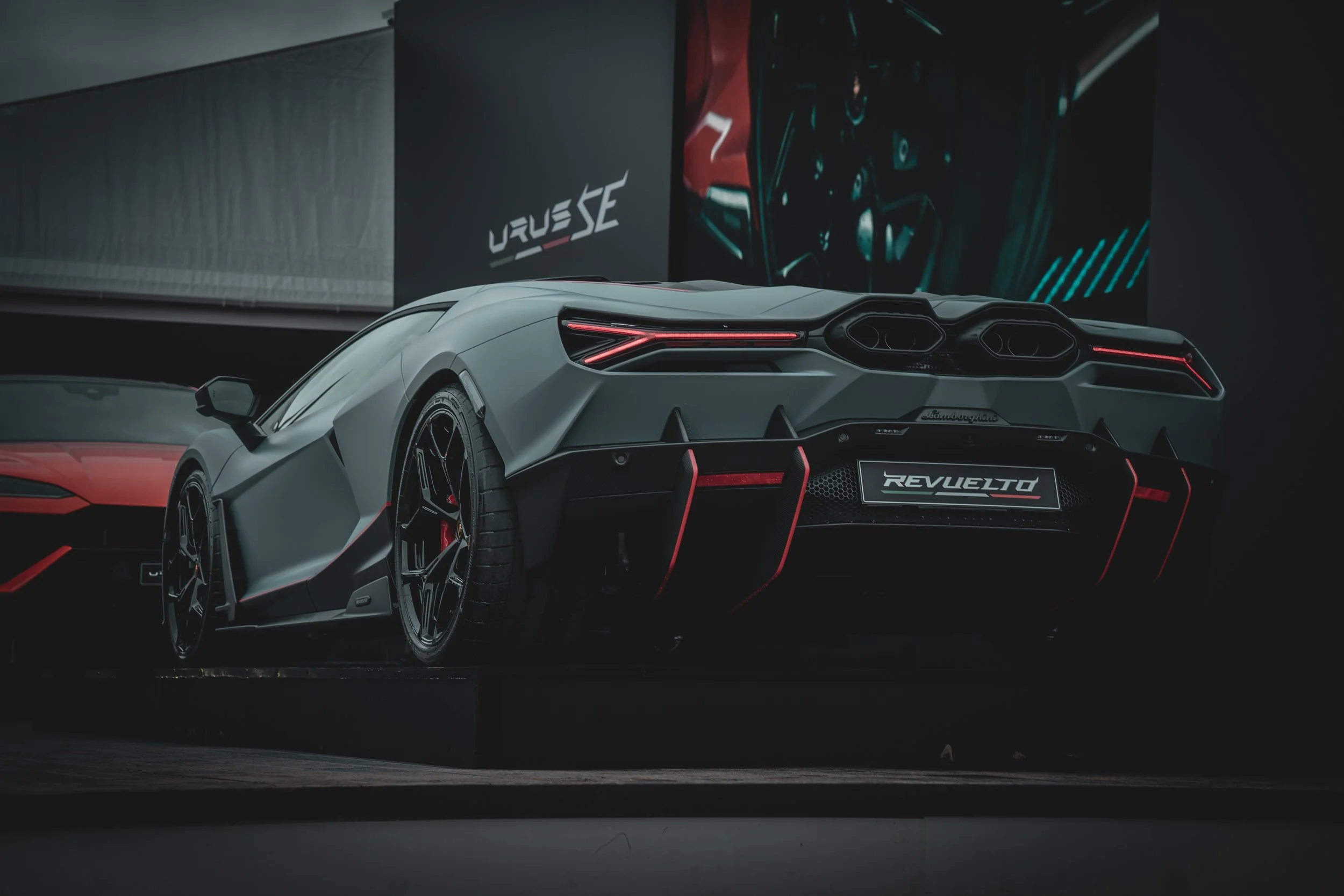 A sleek, black Lamborghini supercar on display at an auto show with the model name 'REVuelto' on the rear plate, and a modern exhibition background featuring the Lamborghini logo and promotional graphics.