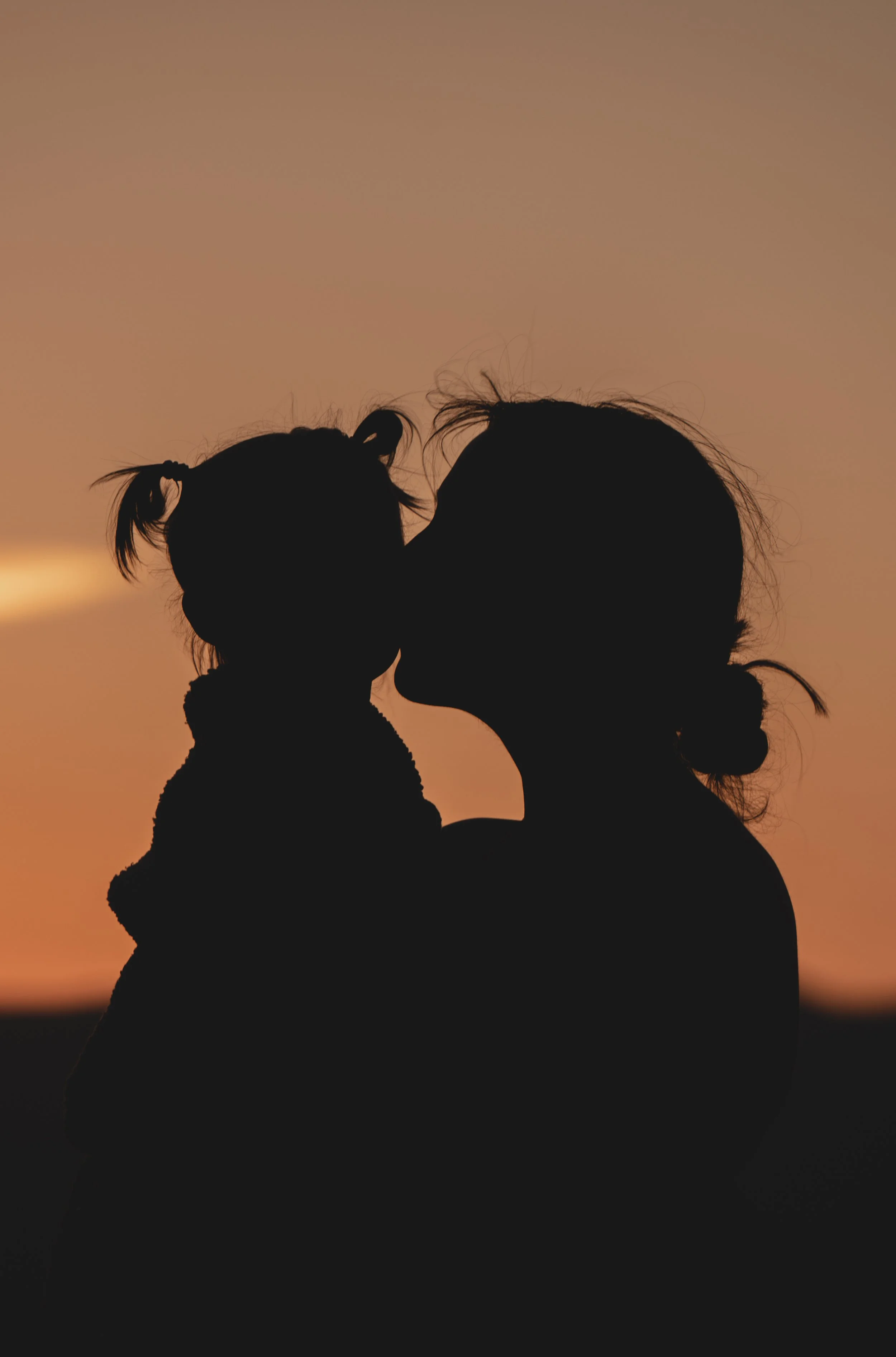 Silhouette of a woman kissing a small child on the cheek during sunset or sunrise.