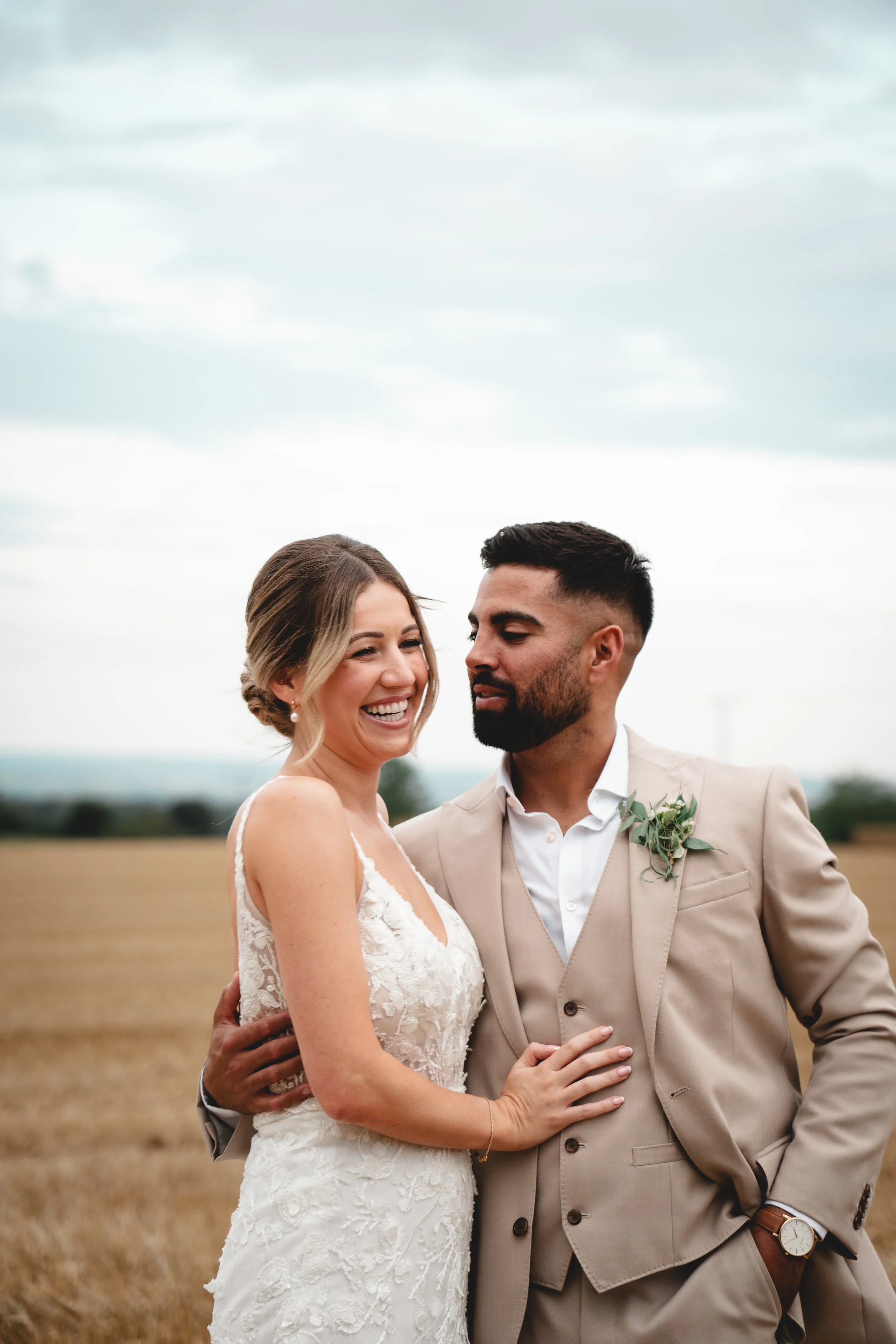 A happy bride and groom on their wedding day in a field, embracing and smiling, with overcast sky in the background.