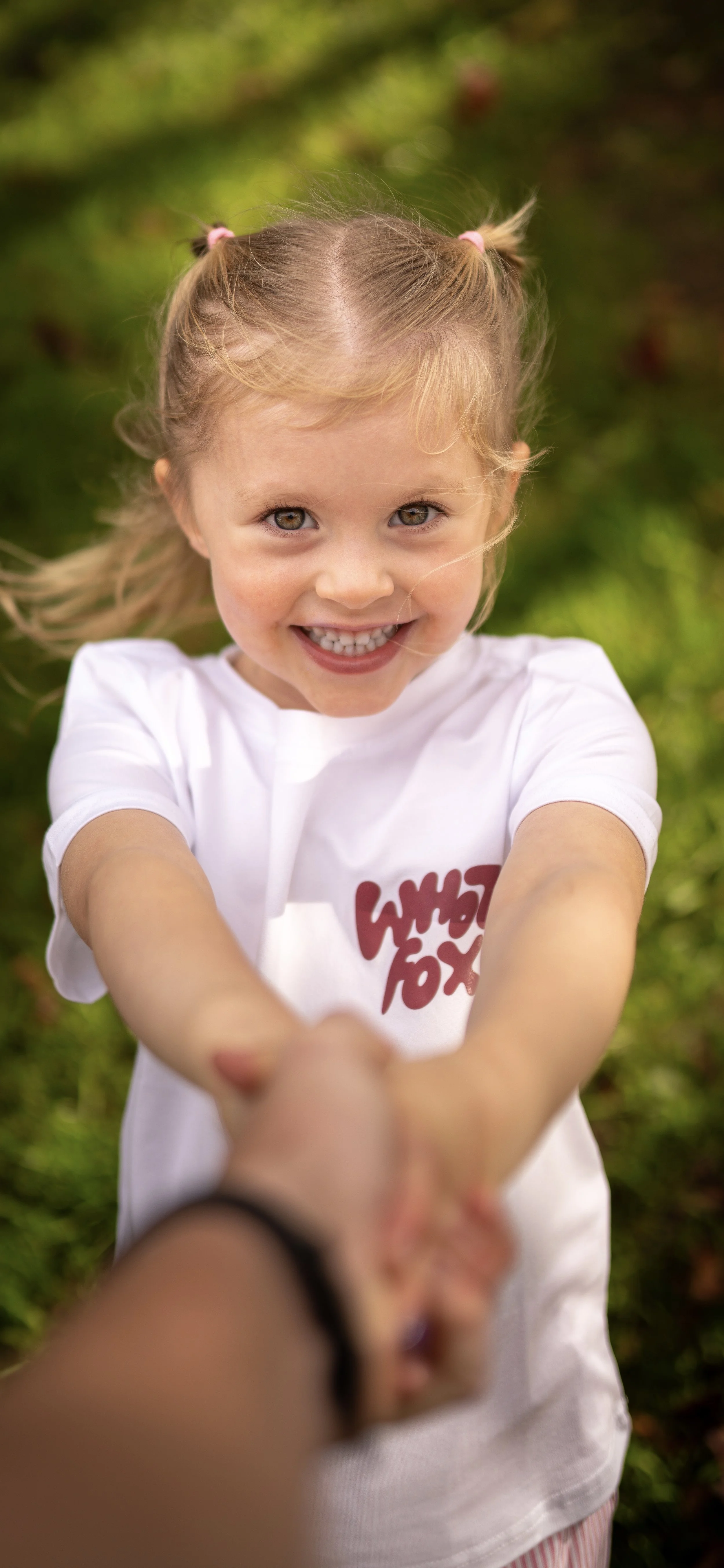 A young girl with blonde hair in pigtails, smiling and holding hands with someone, outdoors on a sunny day with green foliage in the background.