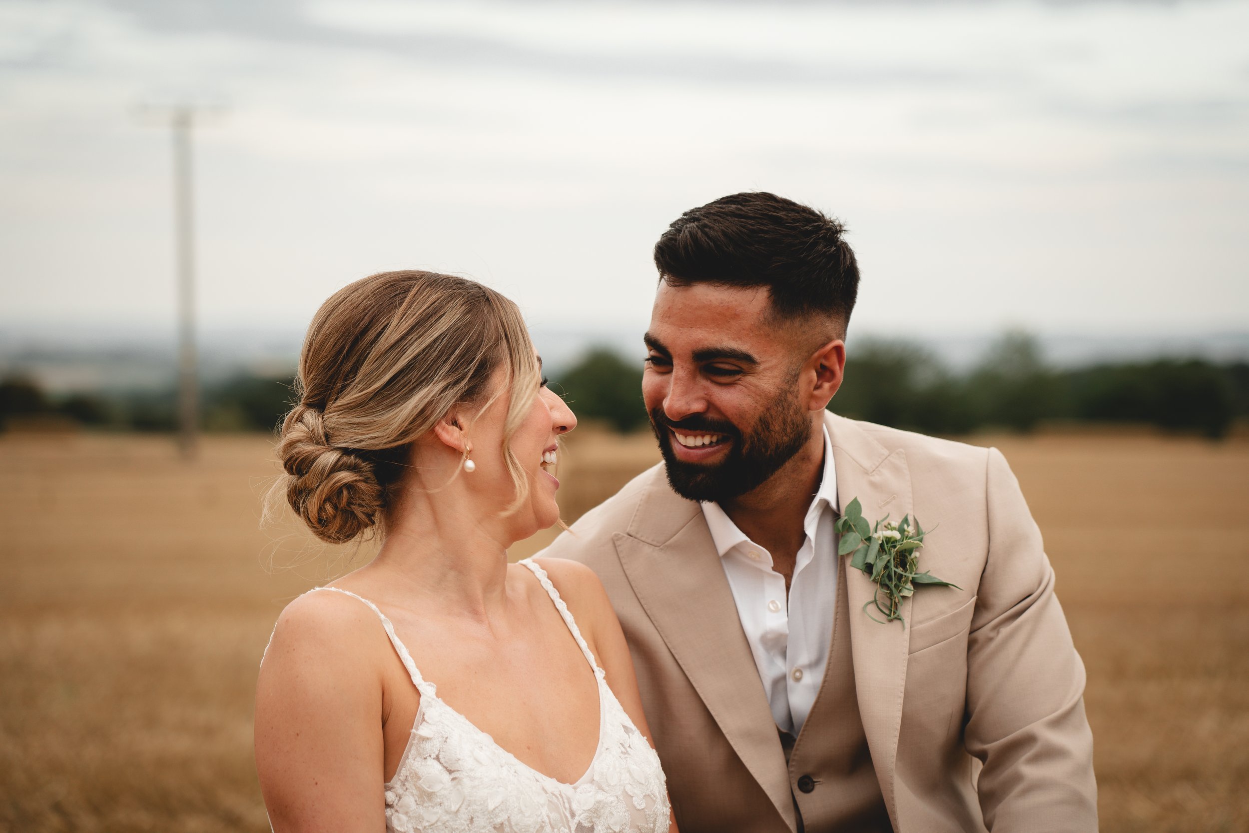 A happy couple outdoors, dressed in wedding attire, smiling and looking at each other in a field with a cloudy sky.