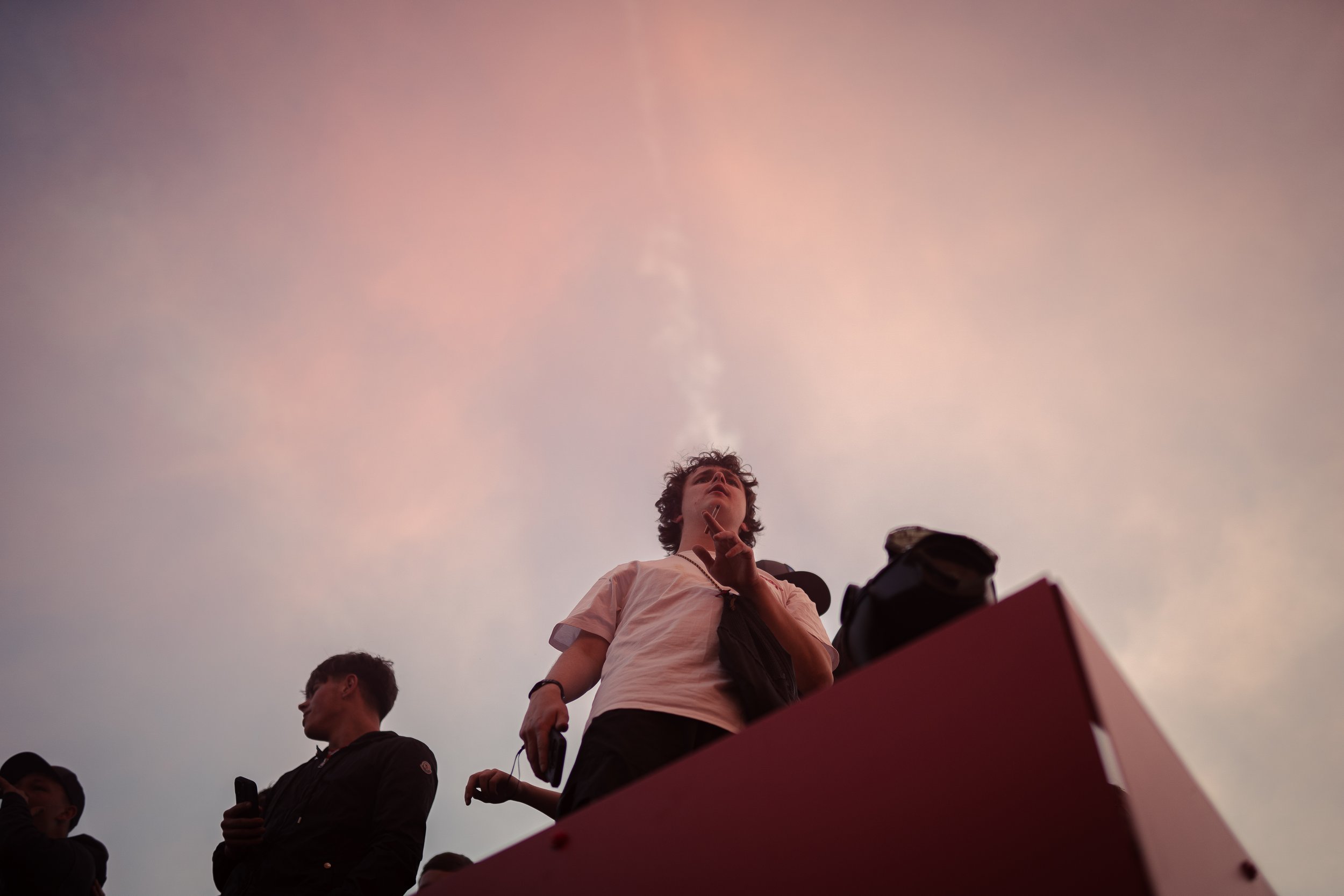 People standing outdoors, with one person smoking and others holding phones, against a sunset or sunrise sky.