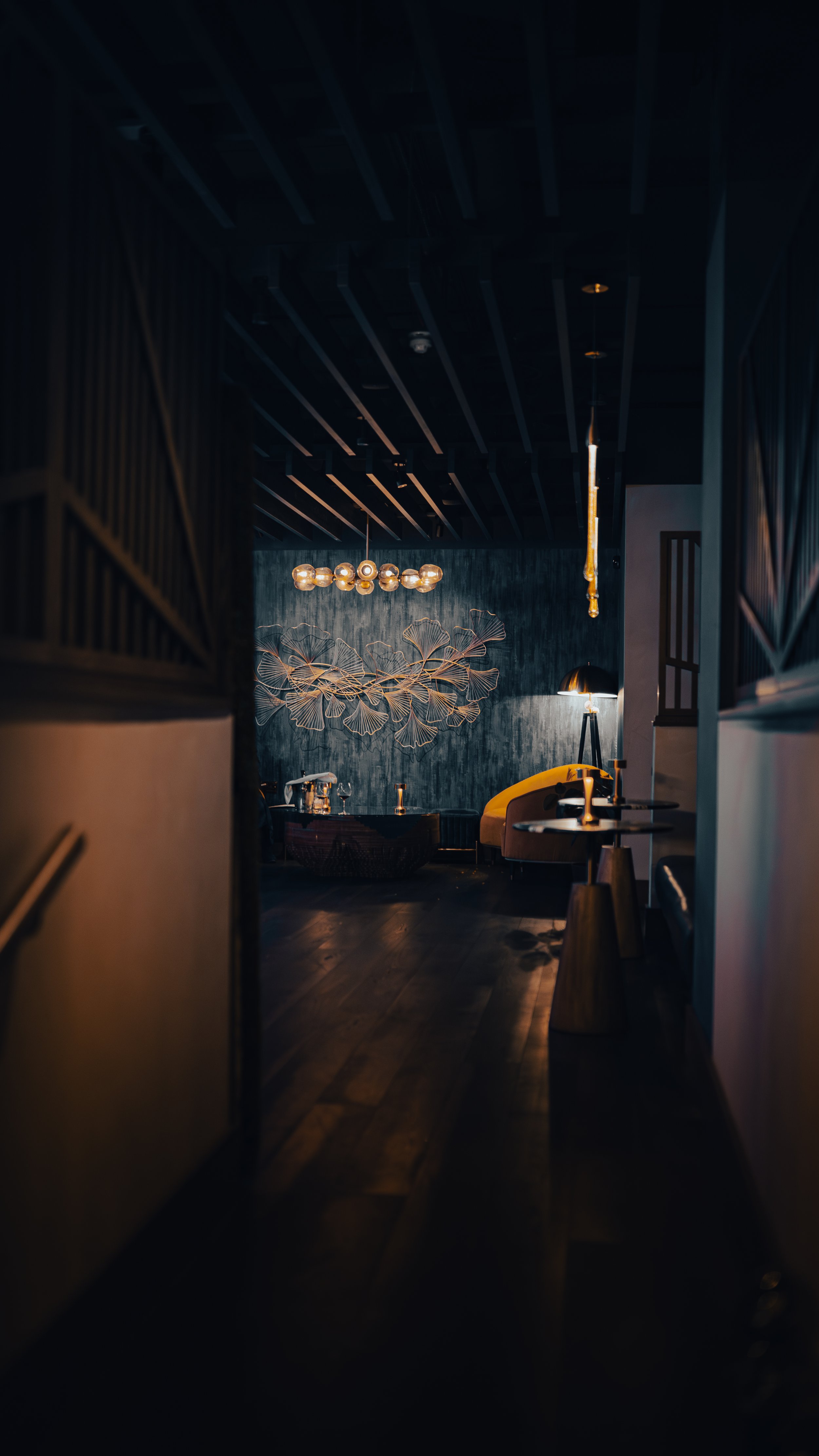 A dimly lit modern lounge with dark walls and wooden flooring, decorated with brass and black lighting fixtures and a yellow sofa.