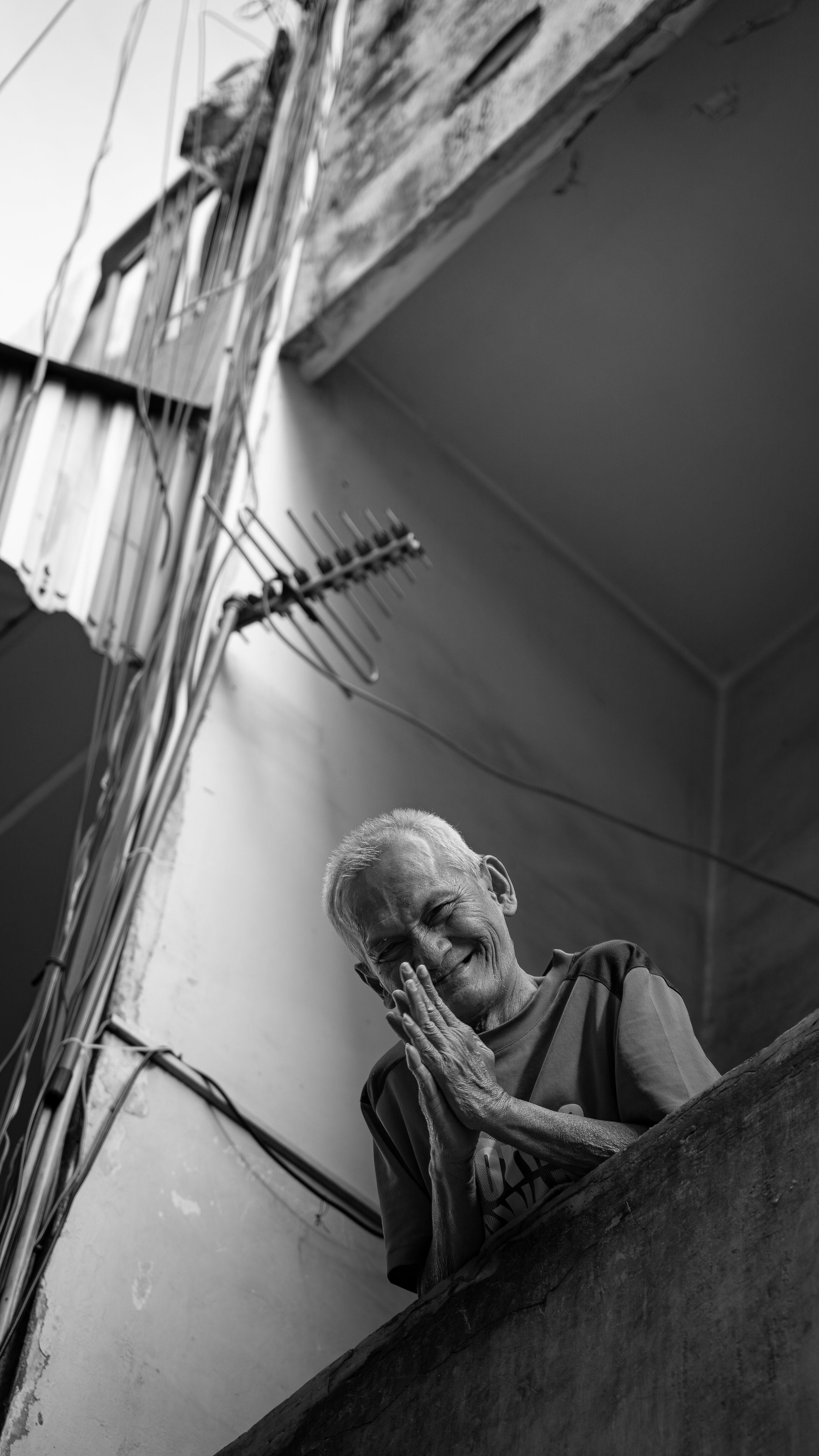 An elderly woman smiling and holding her face, standing on a staircase in a home with exposed wires and a television in the background.