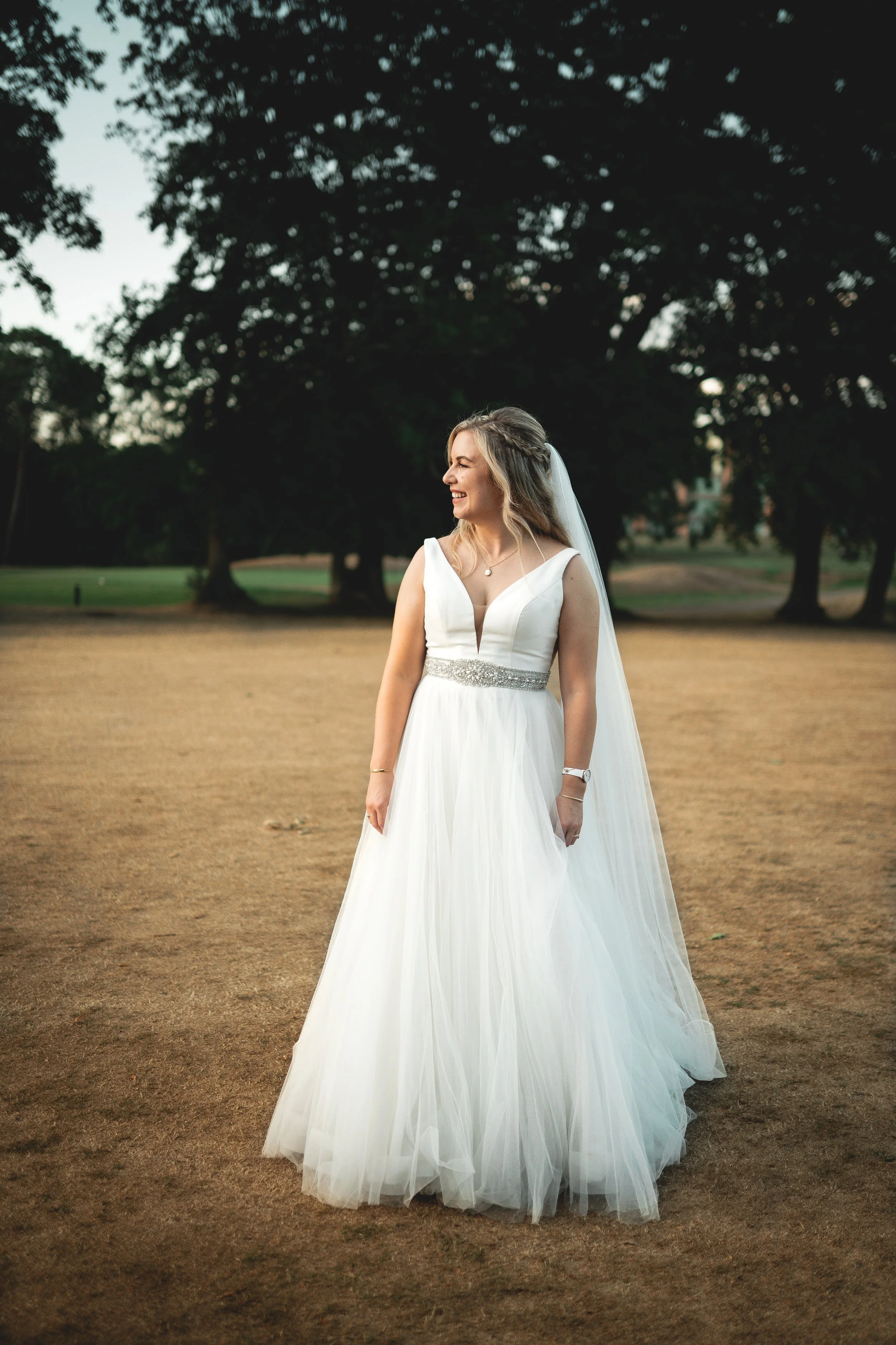 A bride in a white wedding dress standing outdoors on a field with trees in the background, smiling and looking to the side.