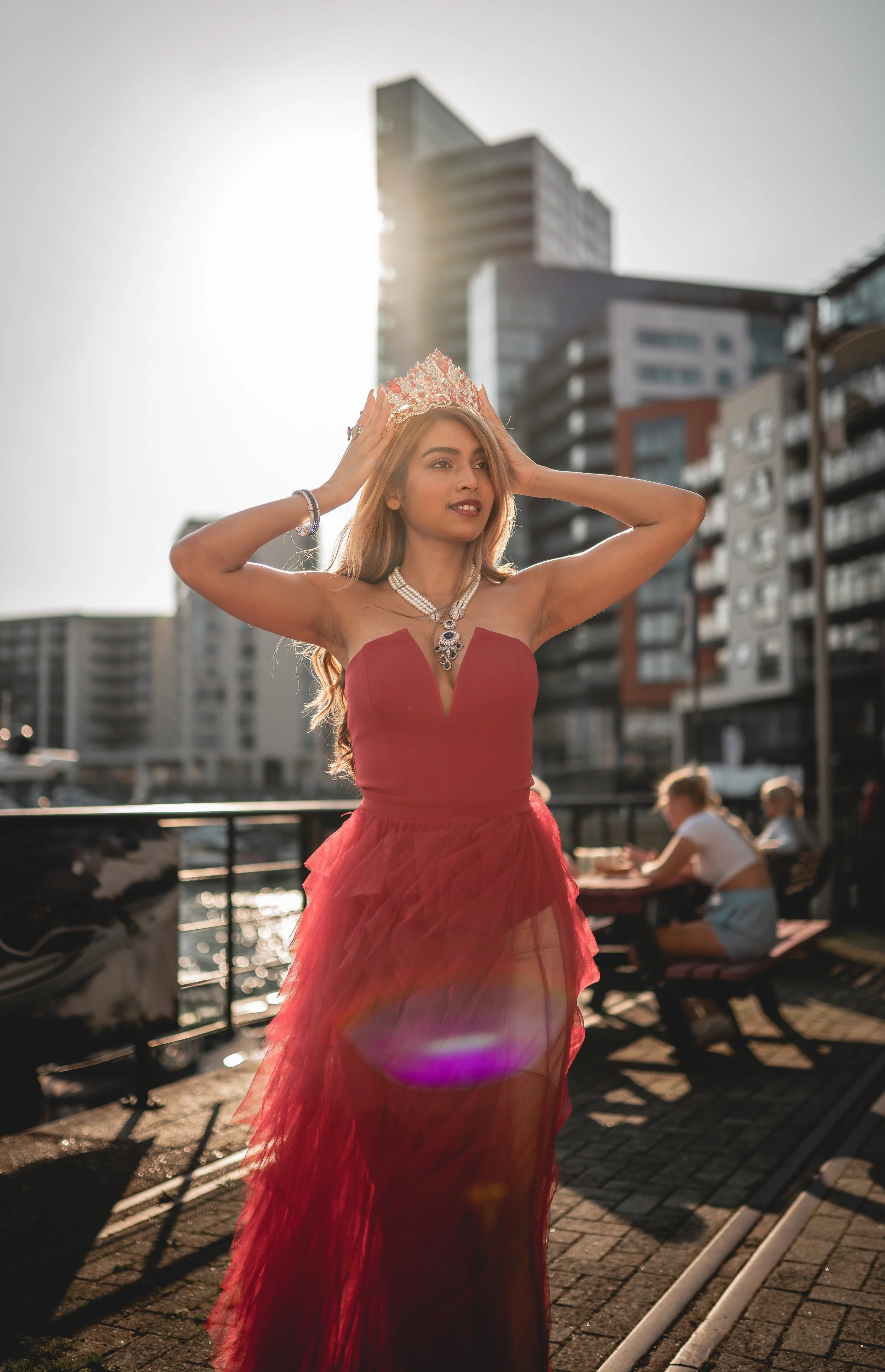 A woman in a red dress and tiara standing outdoors on a sunny day, with tall buildings in the background. She's wearing jewelry and holding her head, possibly adjusting her tiara.