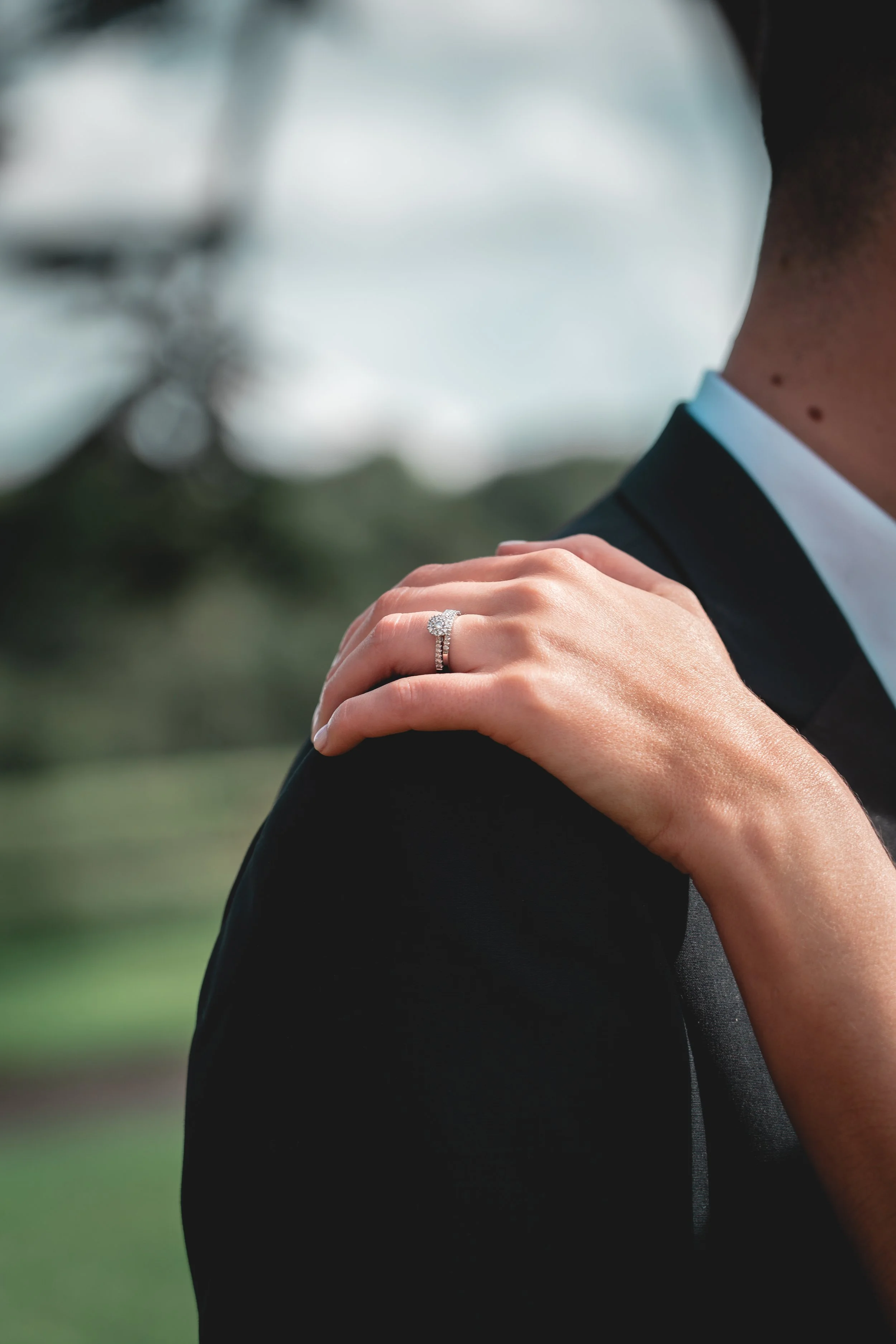 Close-up of a bride's hand with an engagement ring on her finger, resting on a groom's shoulder, with a blurred outdoor background.