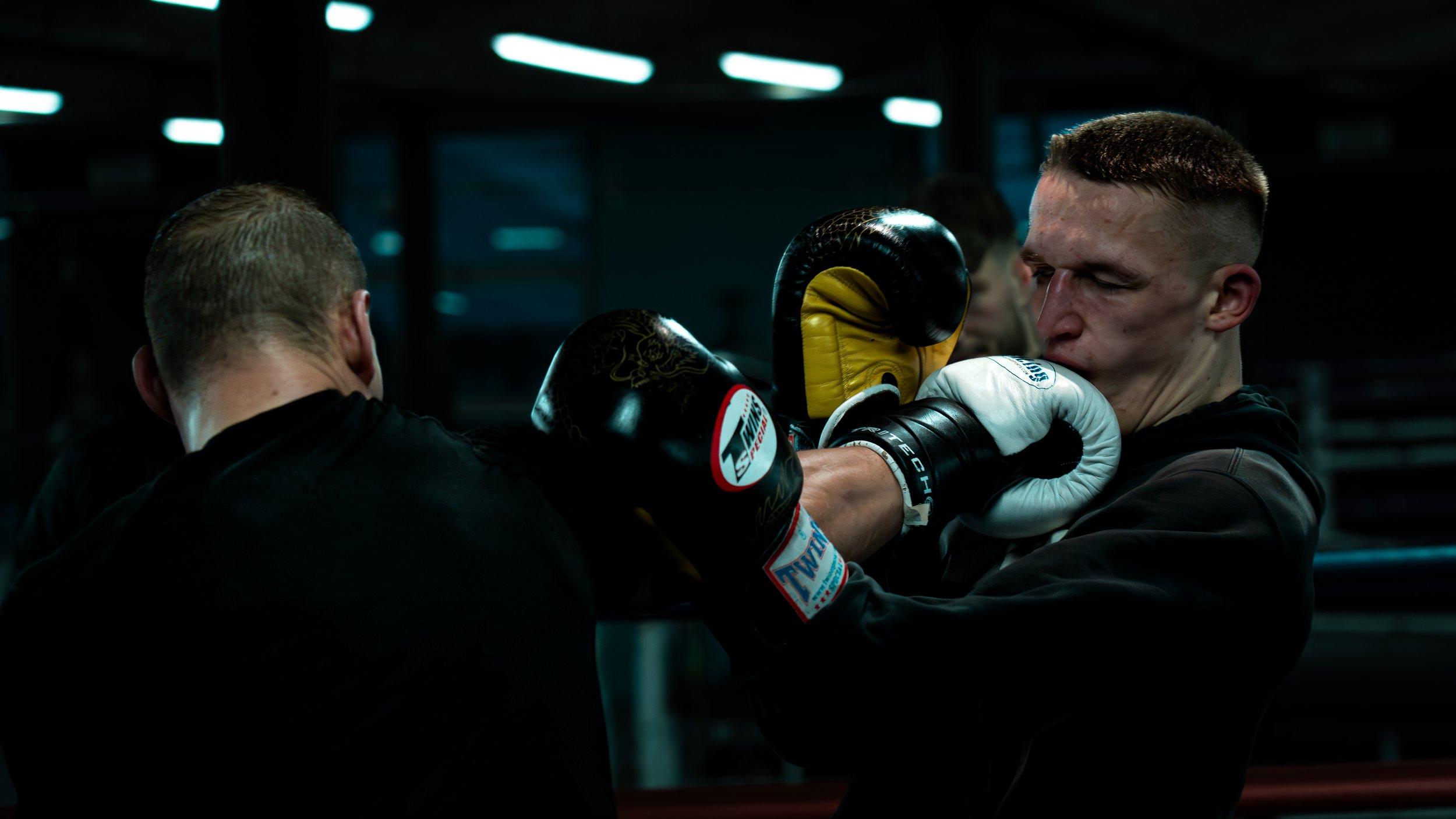 Two men practicing boxing in a gym; one delivering a punch and the other defending with gloves.