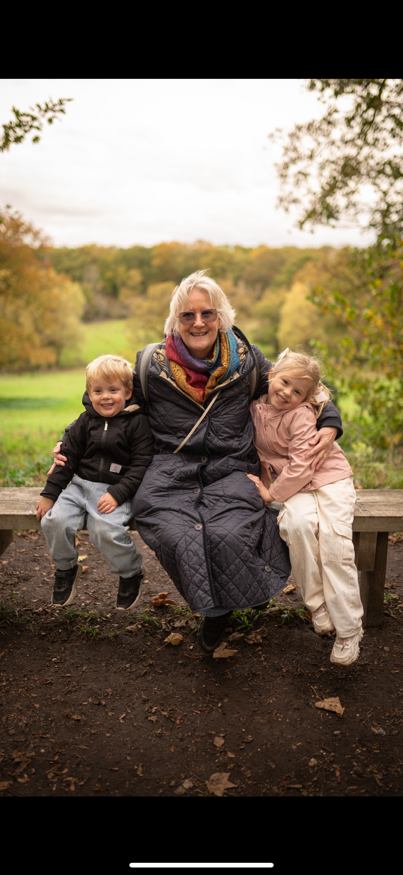 An elderly woman with glasses, sitting on a wooden bench outdoors during fall, smiling with two young children, a boy and a girl, sitting beside her. All are dressed in casual clothes suitable for cool weather, with autumn-colored trees in the backgr